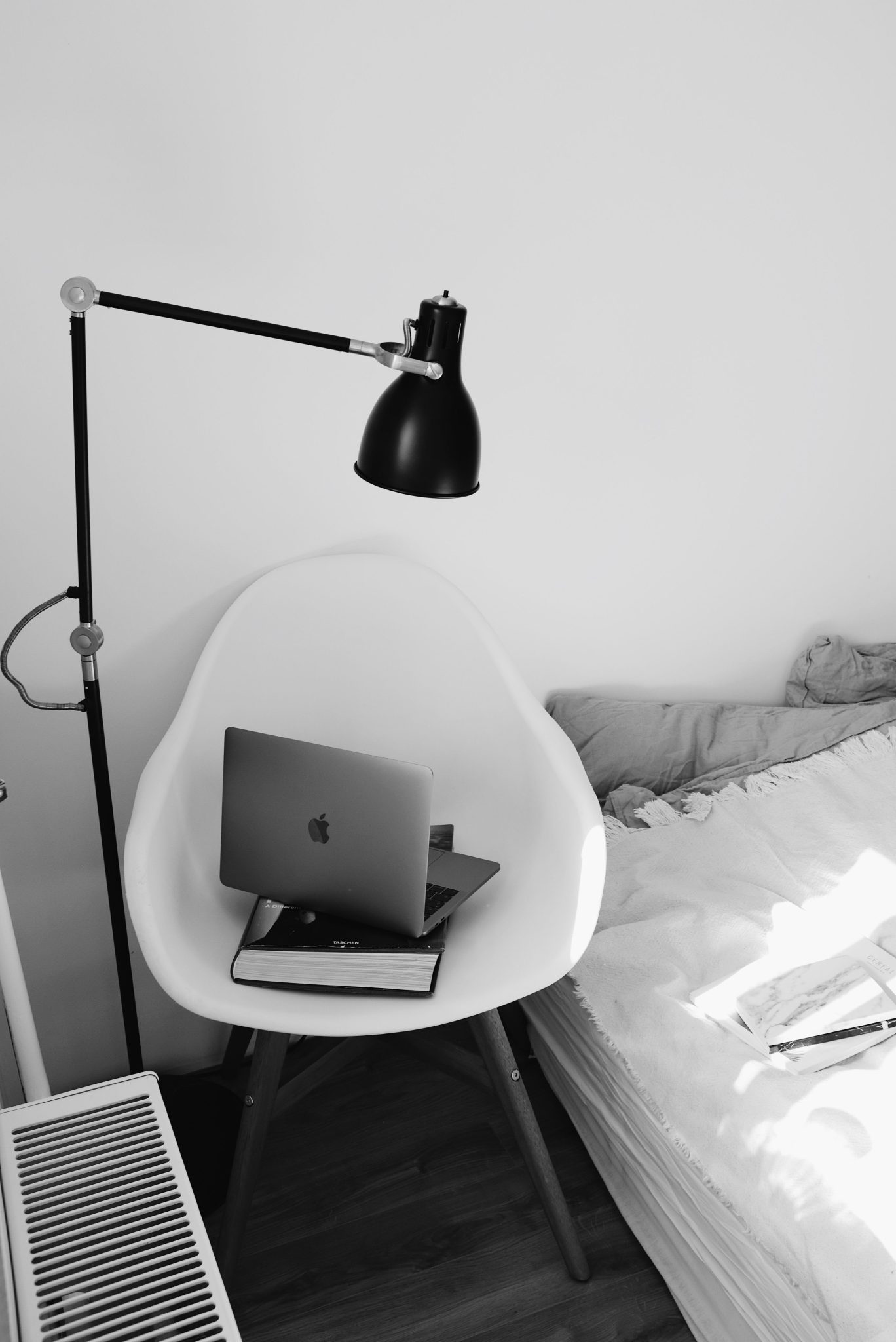 Black and white modern interior of workplace with laptop placed on big book on white chair under lamp near bed in light modern bedroom in apartment