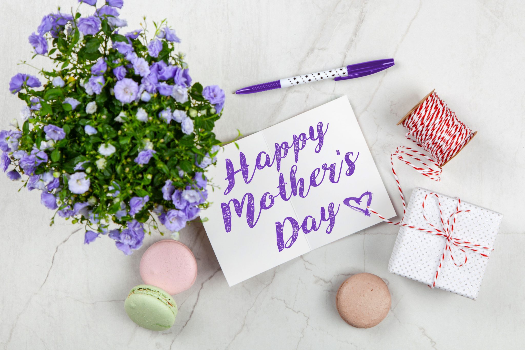 Flat lay of Mother's Day card with purple flowers, gift, macarons, and a pen indoors.