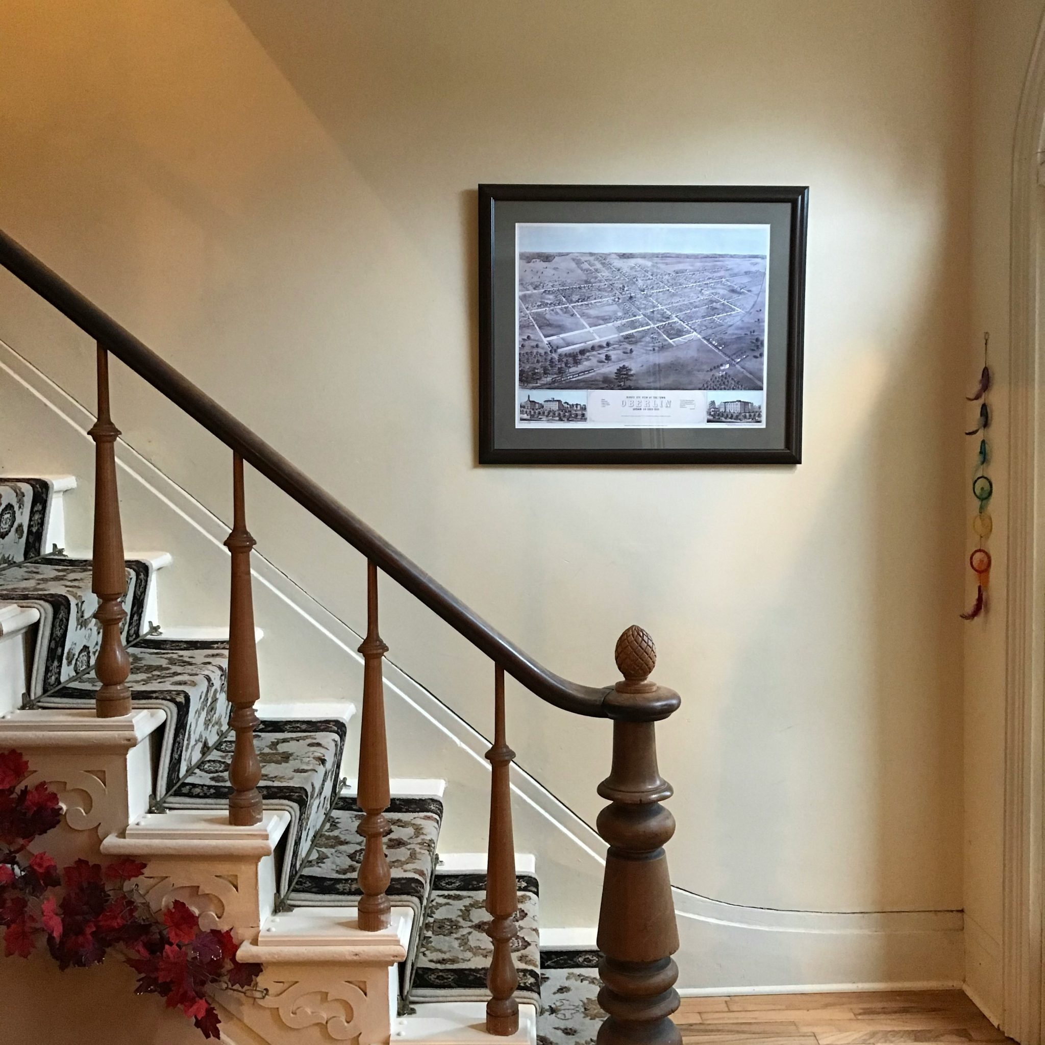 Staircase with s patterned runner in a 150-year-old country home. The walls are cream-colored. The stairs are white. The bannister is hand-carved. A vintage map hangs framed upon the wall.