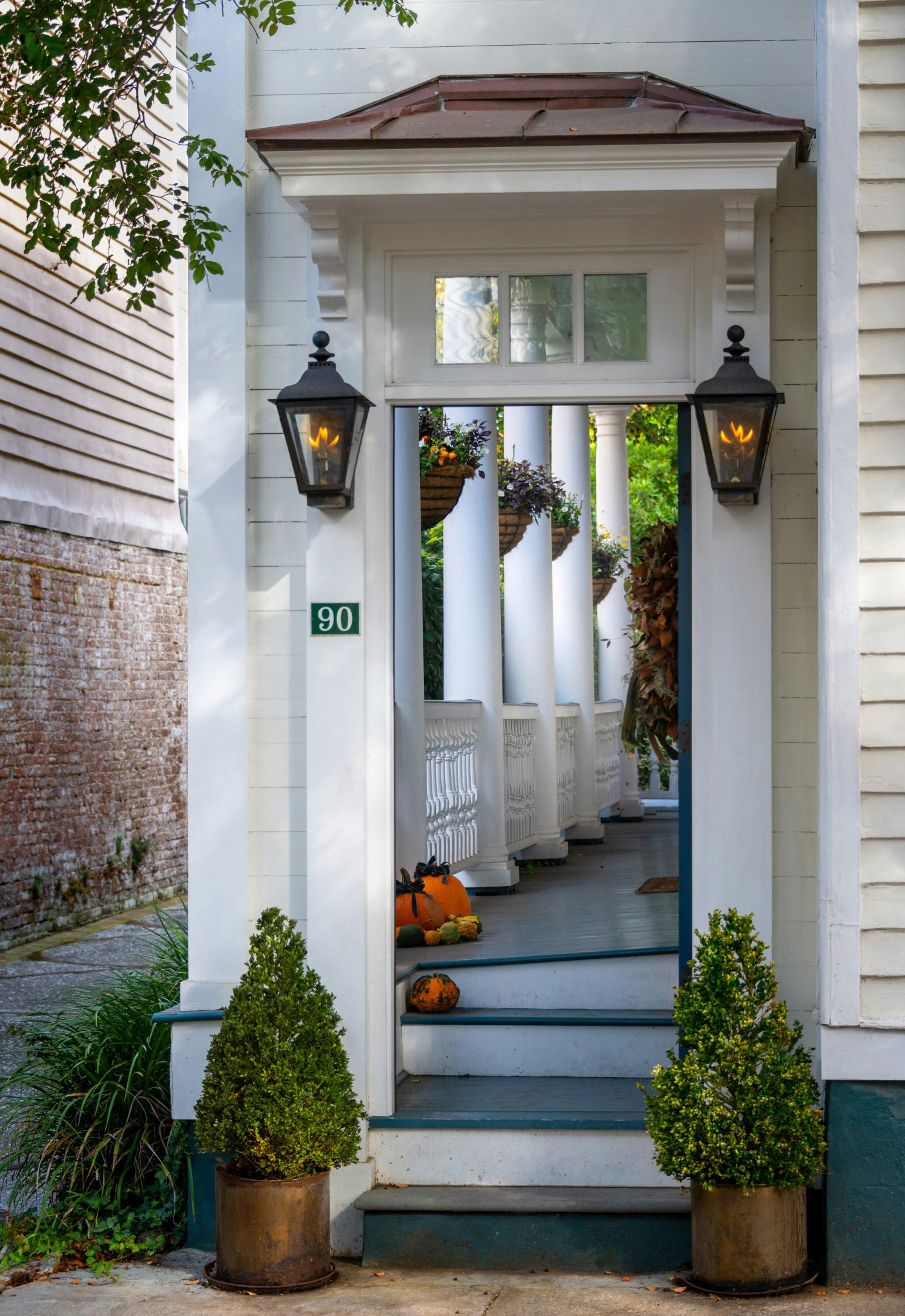 A charming Charleston porch framed by gas lanterns, adorned with pumpkins and lush hanging plants, evokes classic Southern elegance in autumn.
