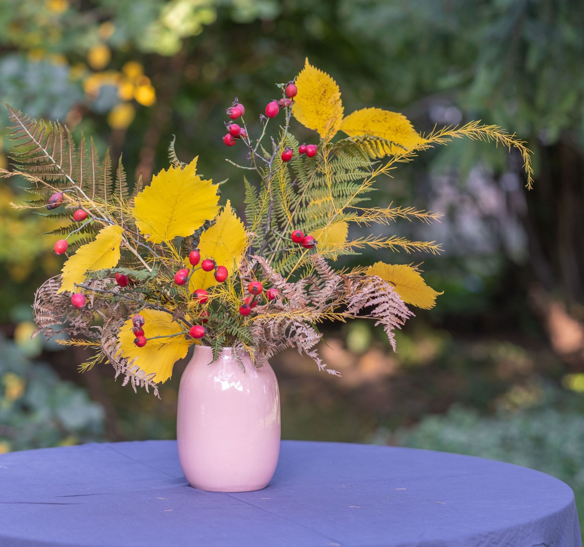 A colorful autumn bouquet with yellow leaves and red berries in a pink vase on a table outdoors.