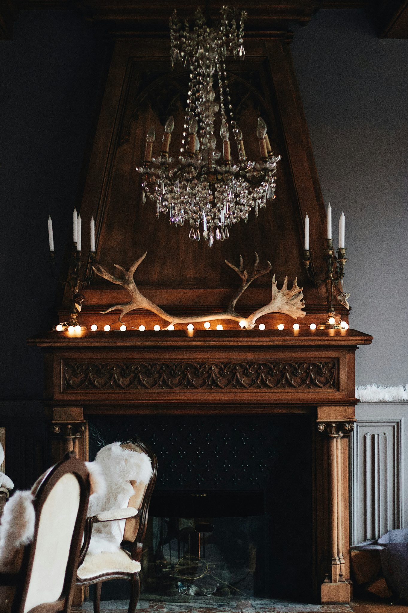 A crystal chandelier above a wooden fireplace mantel decorated with antlers and string lights