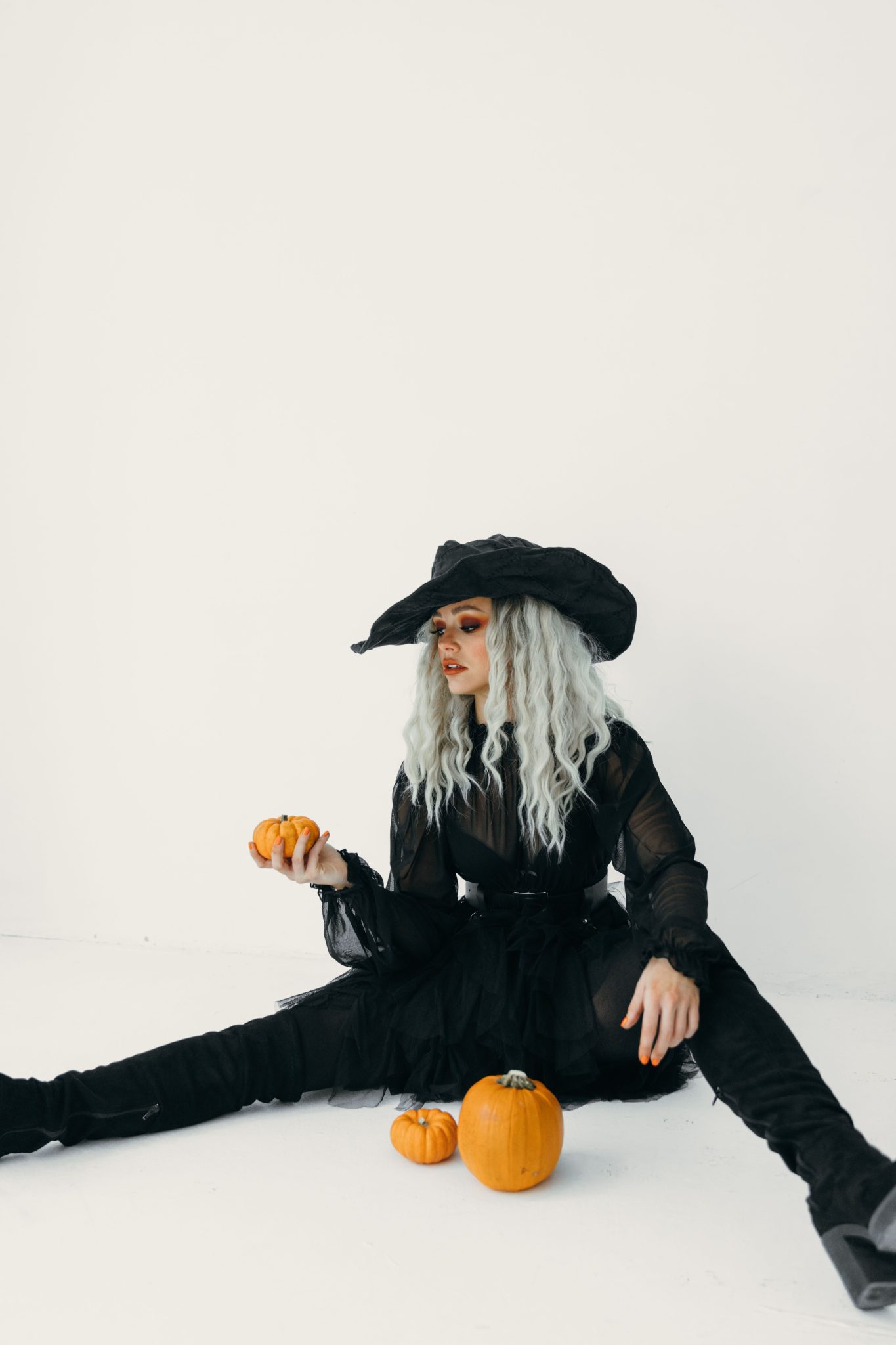A fashionable woman in a witch costume sitting indoors with pumpkins, perfect for Halloween themes.
