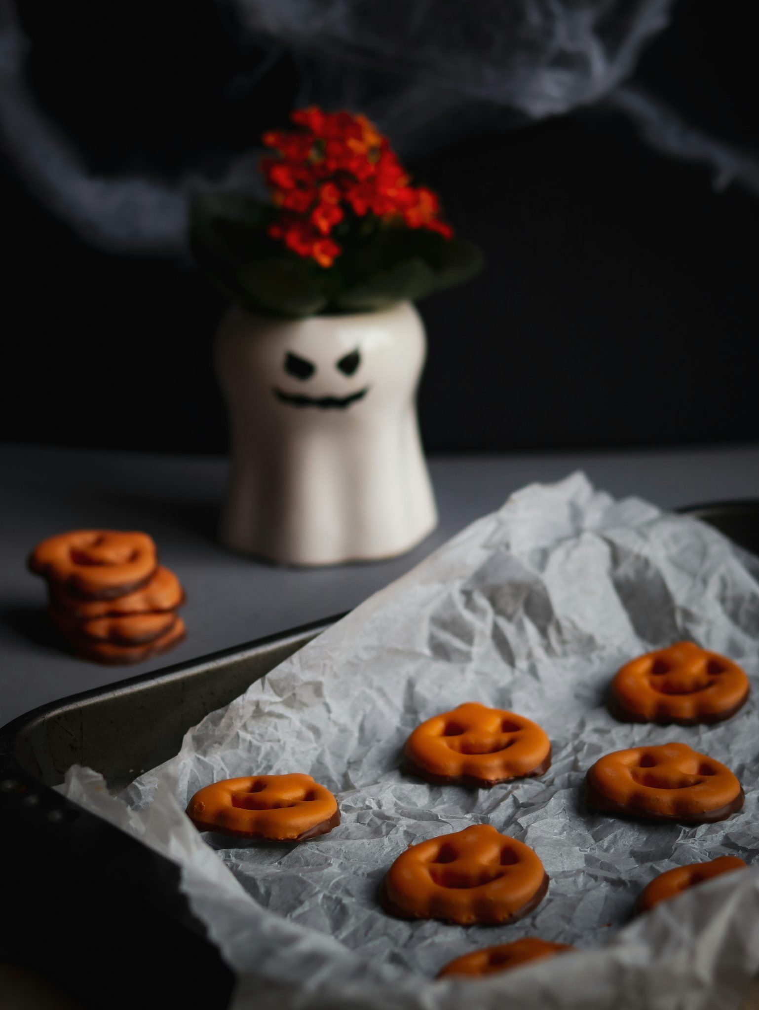 A festive Halloween-themed setup featuring a baking tray of adorable pumpkin-shaped pretzels coated in orange chocolate, giving a sweet and spooky vibe. In the background, a ghost-themed vase with a b