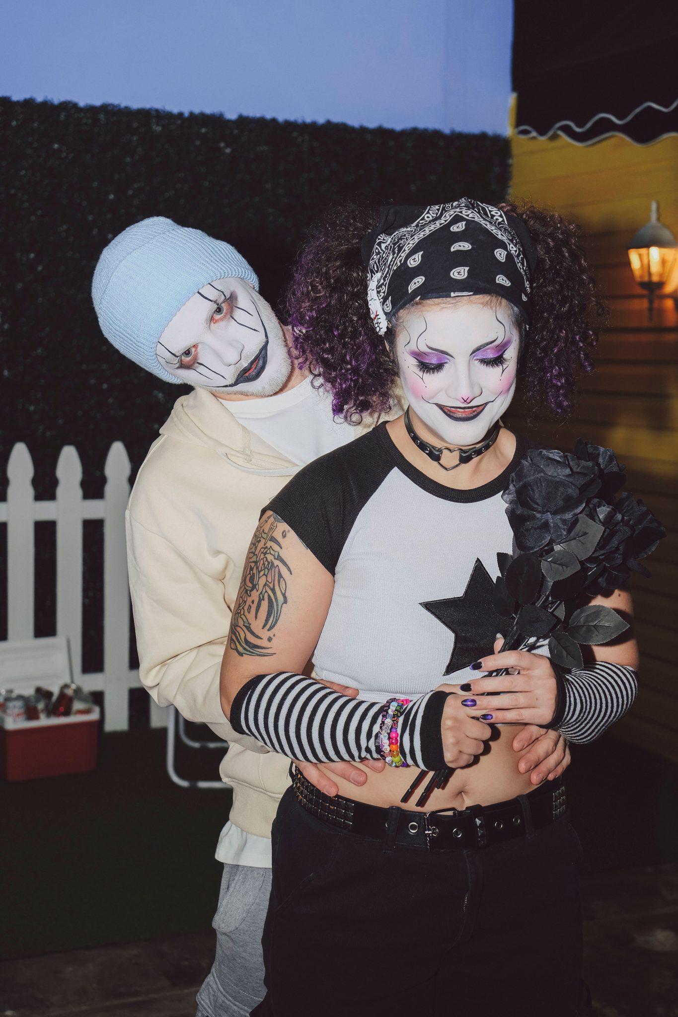 A gothic-styled couple in face paint and costumes share a tender embrace indoors.
