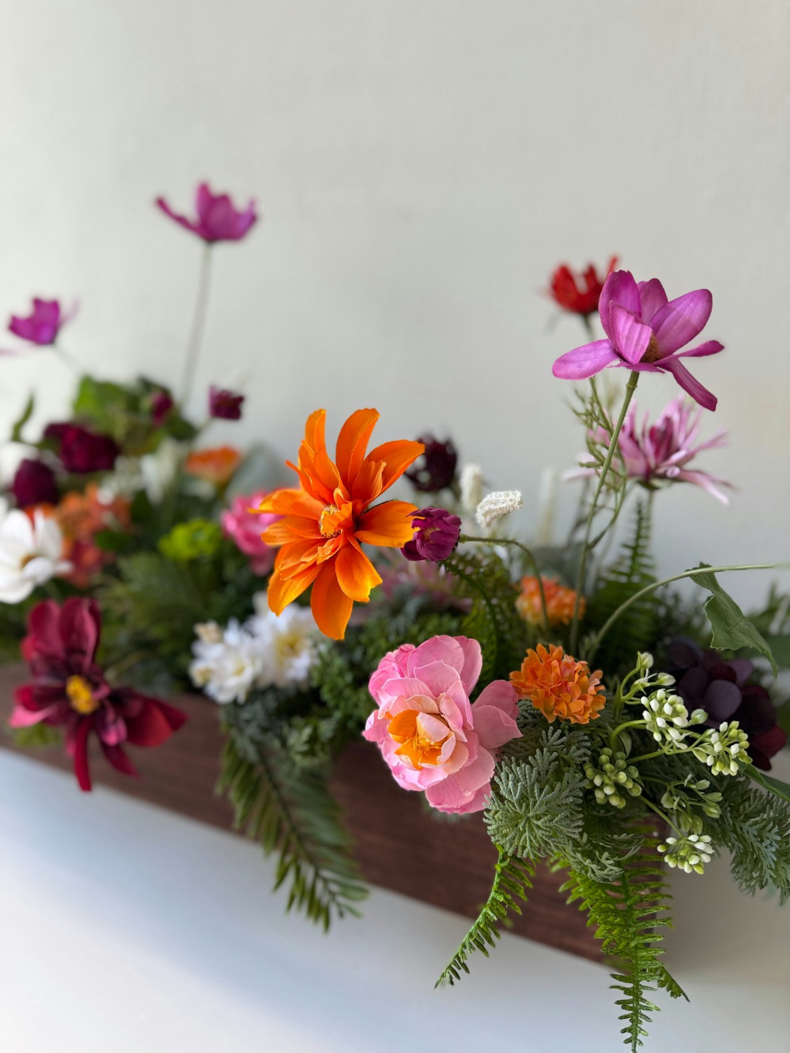A lively, garden-inspired floral arrangement set in a rustic wooden planter, featuring a colorful mix of blooms and greenery. Bright orange cosmos, soft pink ranunculus, magenta and purple flowers, an