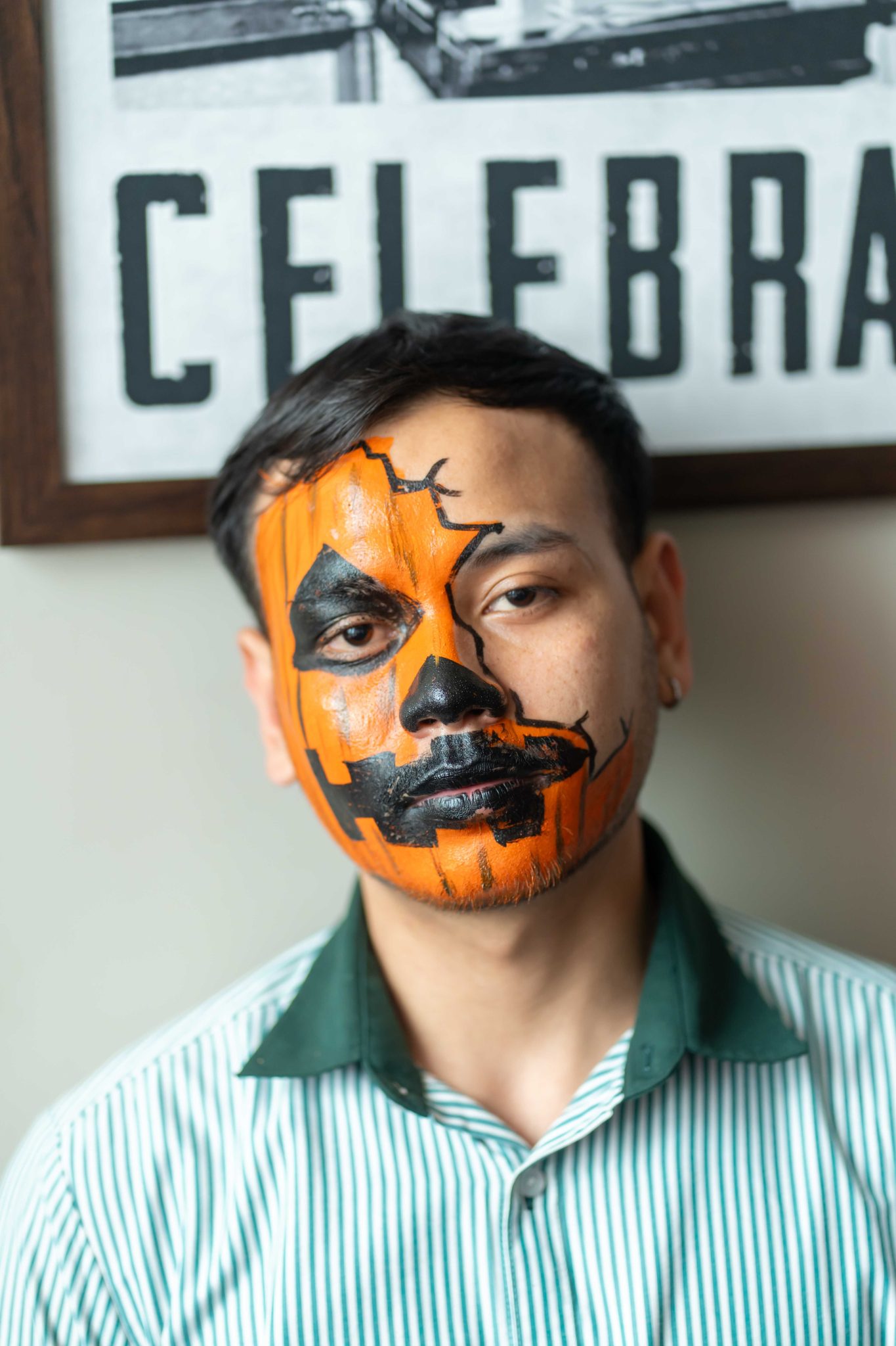 A man with pumpkin-themed face paint poses near a celebration sign, embodying Halloween spirit.