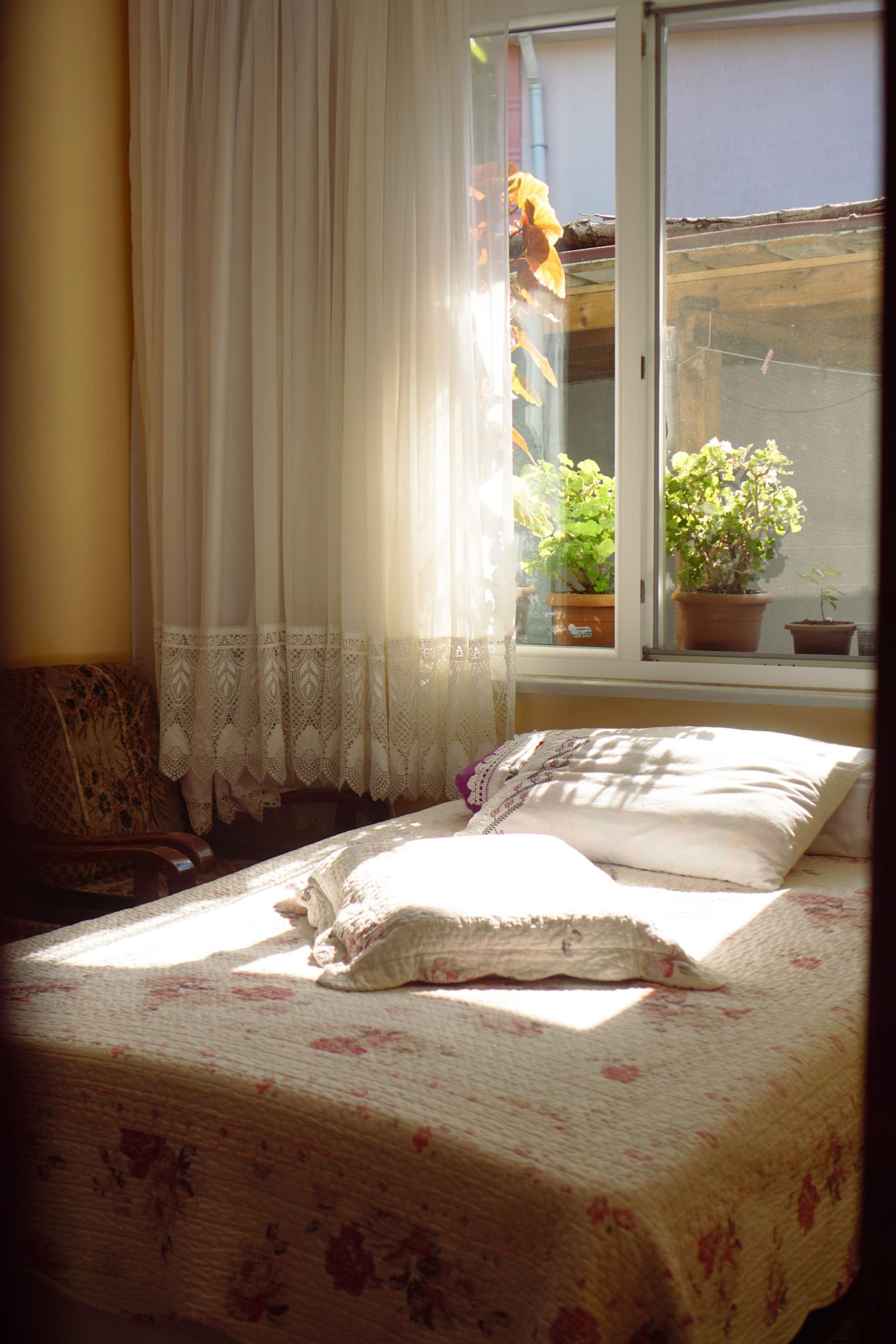 A serene bedroom with sunlight streaming through lace curtains onto a floral bedspread.