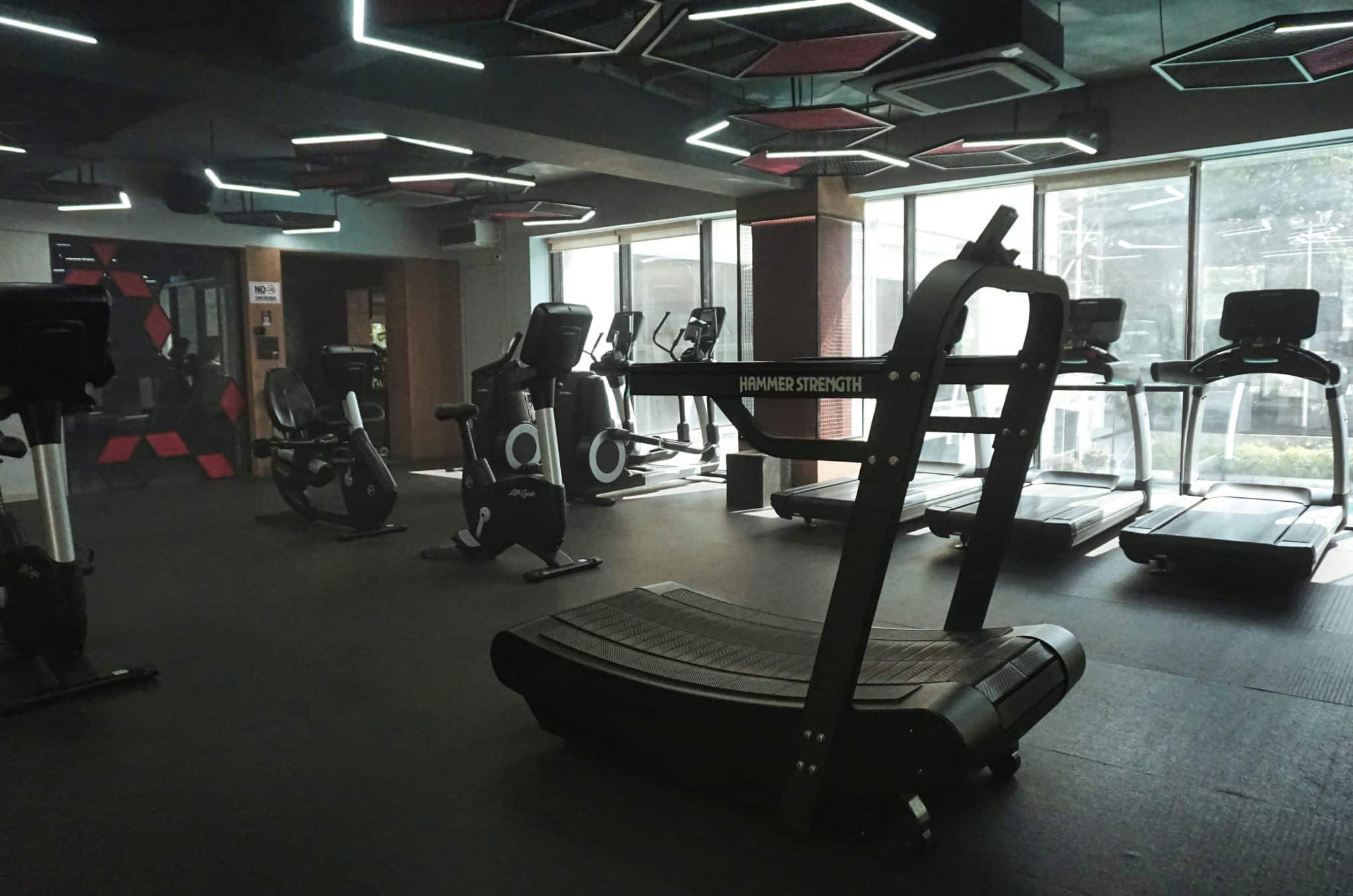 A sleek, modern fitness center featuring high-end cardio machines and strength equipment under distinctive geometric LED lighting. The space combines a professional industrial aesthetic with floor-to-