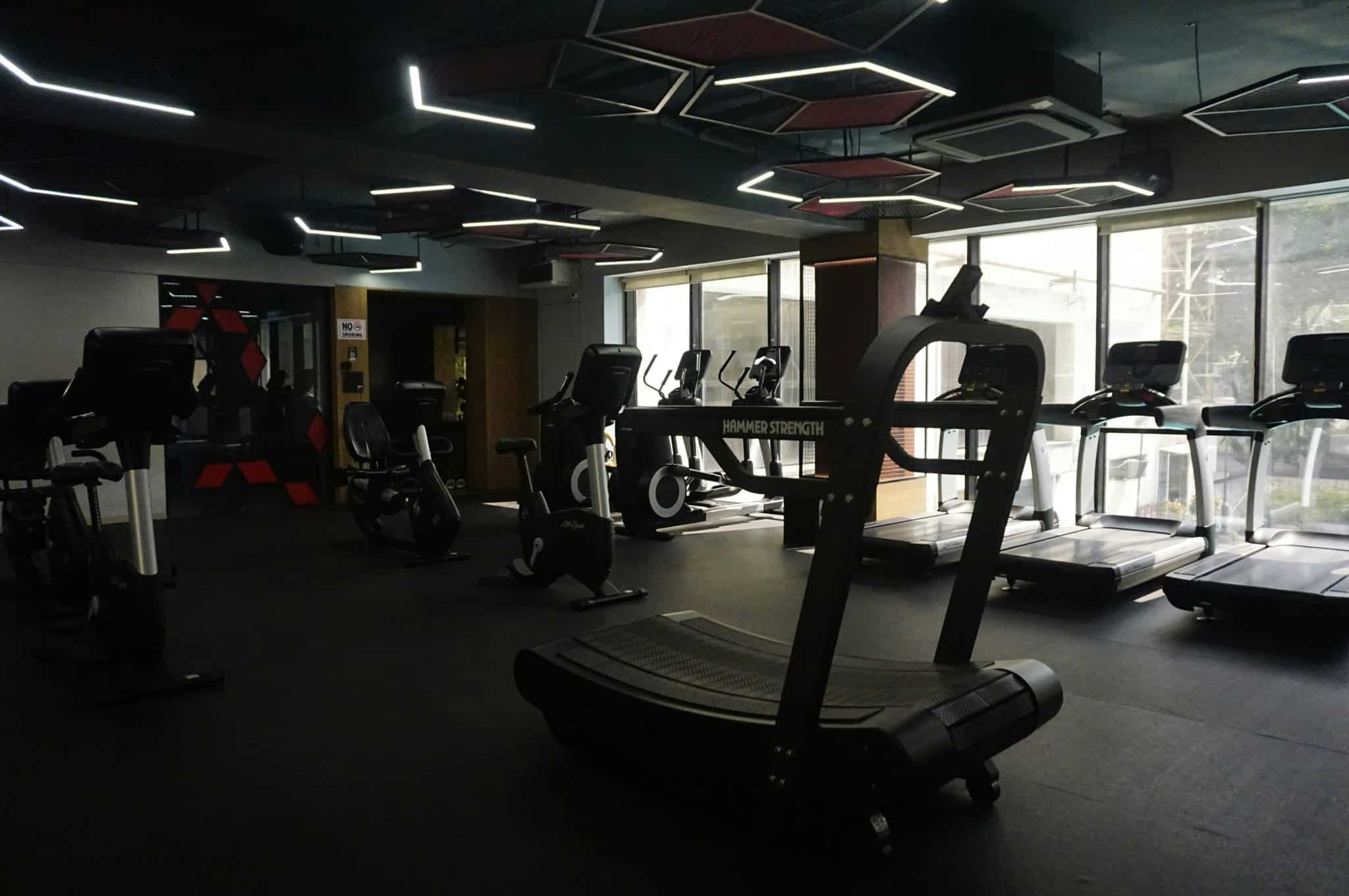 A sleek, modern fitness center featuring high-end cardio machines and strength equipment under distinctive geometric LED lighting. The space combines a professional industrial aesthetic with floor-to-