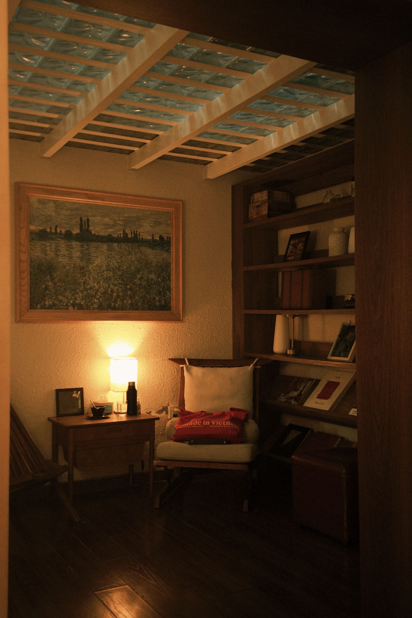 A small, cozy nook featuring a comfortable chair, softly lit lamp, and wooden bookshelf filled with books.