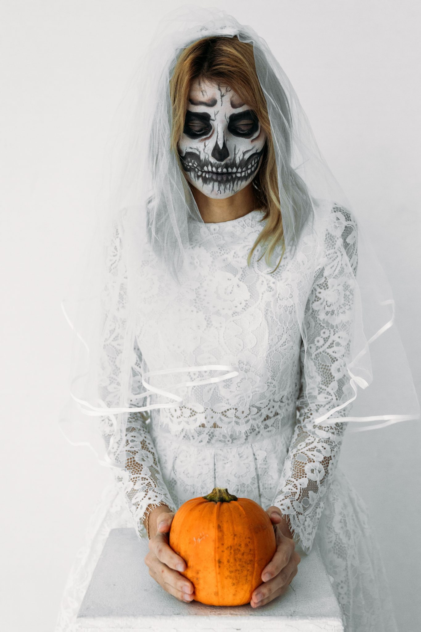 A spooky Halloween bride in a white lace dress with skull face paint holding a pumpkin.