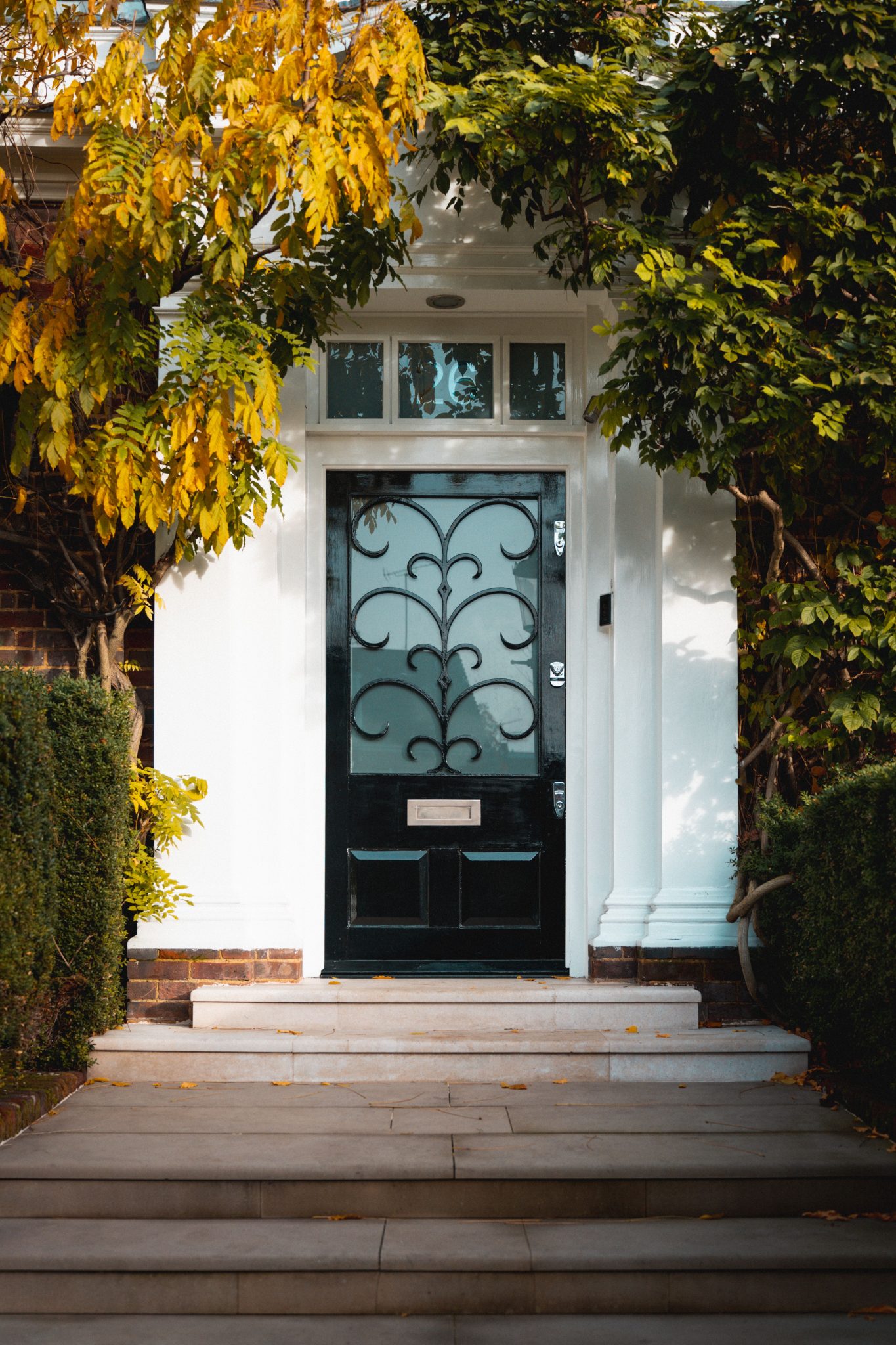 A stylish black door adorned with decorative elements surrounded by autumn foliage in a London neighborhood.