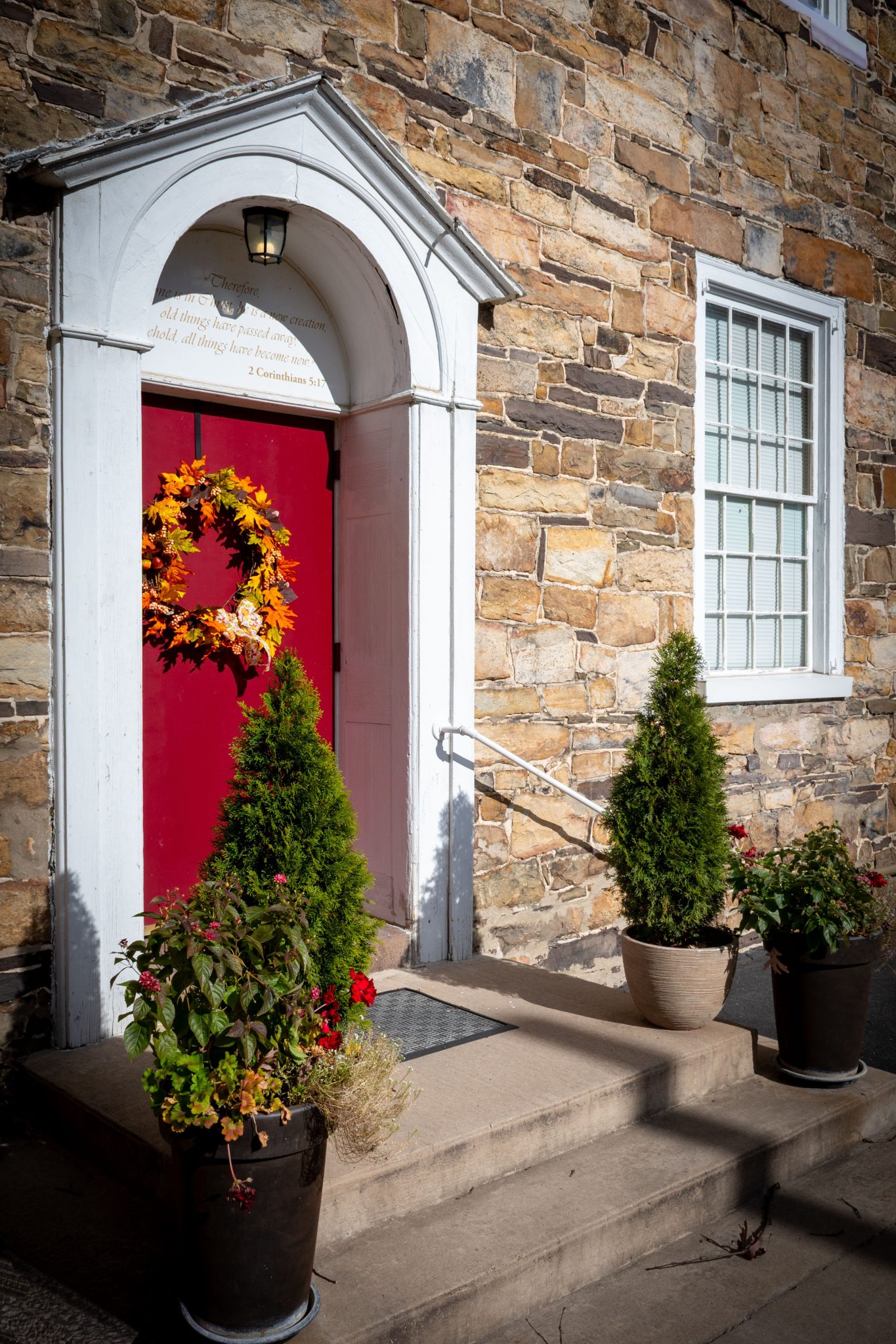 A vibrant red door with fall wreath and potted plants on stone building.