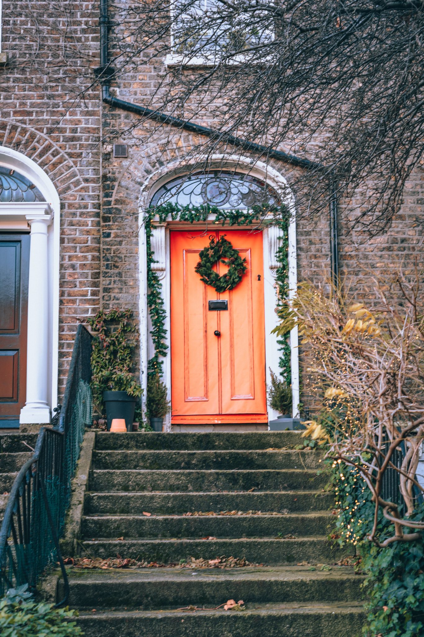A vintage brick entrance featuring a bold red door decorated with a festive wreath, framed by greenery.
