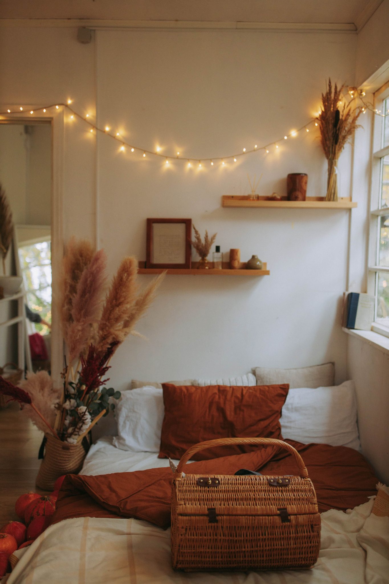 A warm and inviting bedroom interior featuring a wicker basket, fairy lights, and pampas grass.