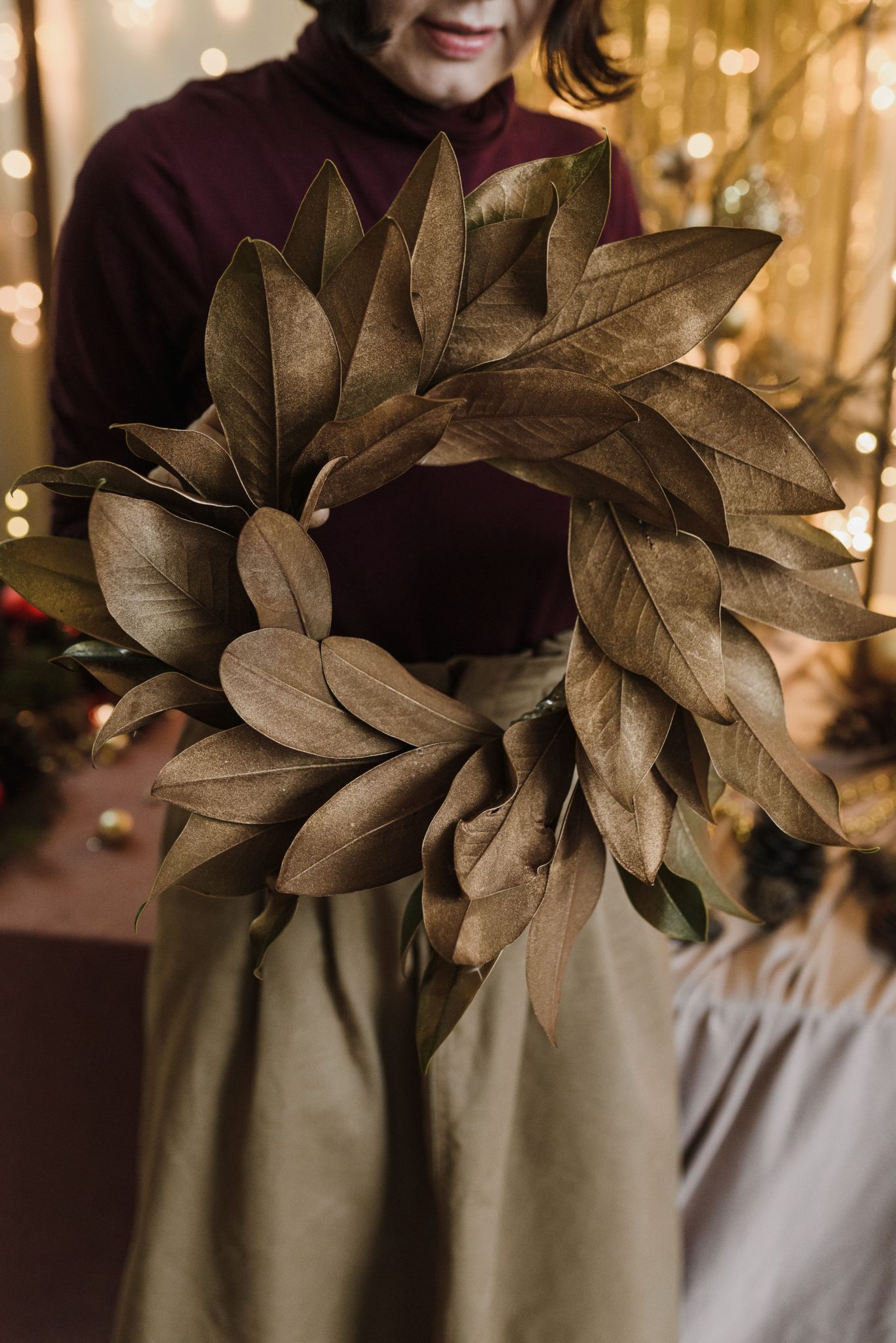 A woman in a turtleneck holds a handmade brown leaf wreath indoors, capturing a warm festive atmosphere.