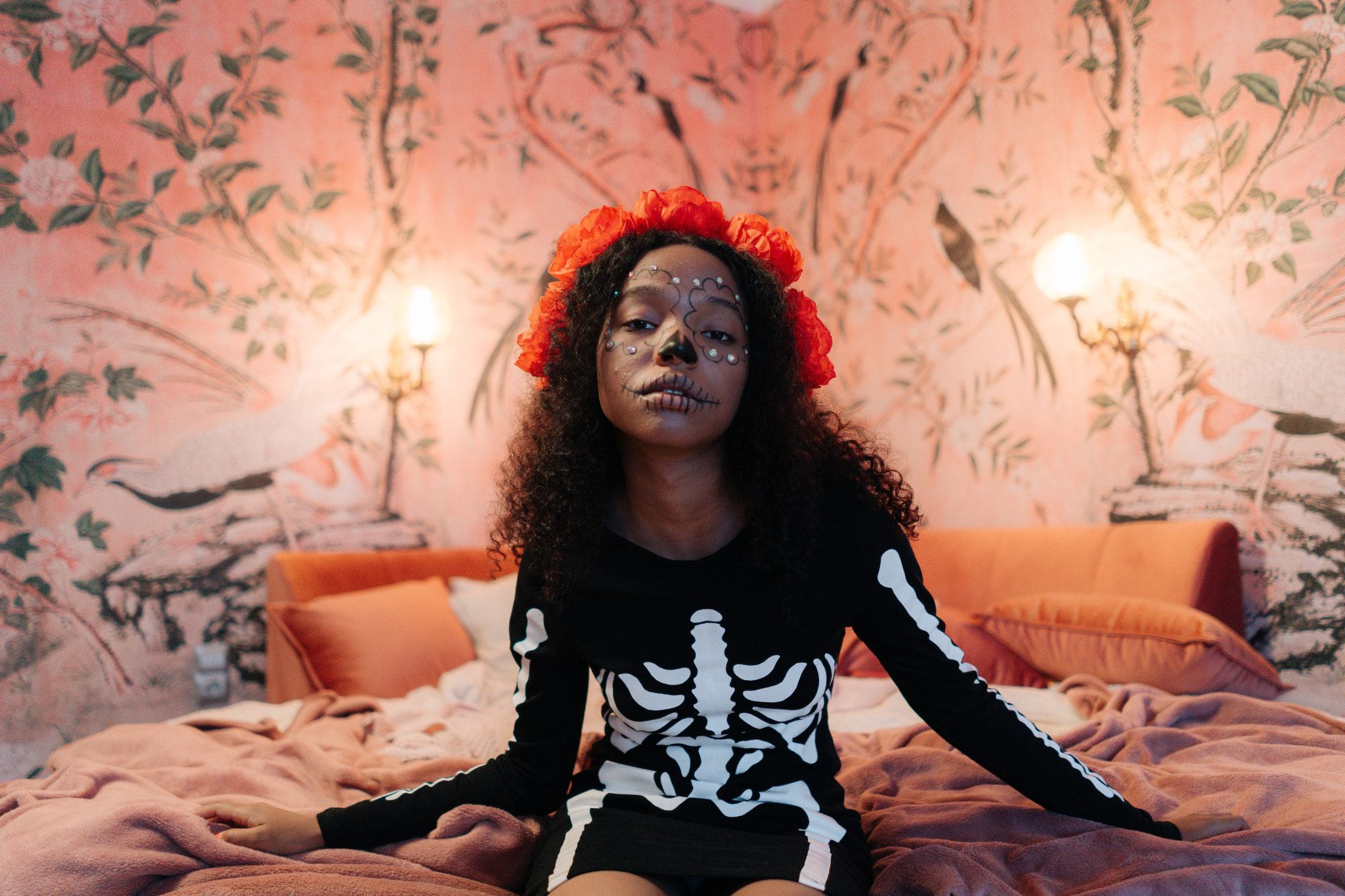 A young girl in a skeleton costume sits on a bed, adorned with face paint and a floral wreath.