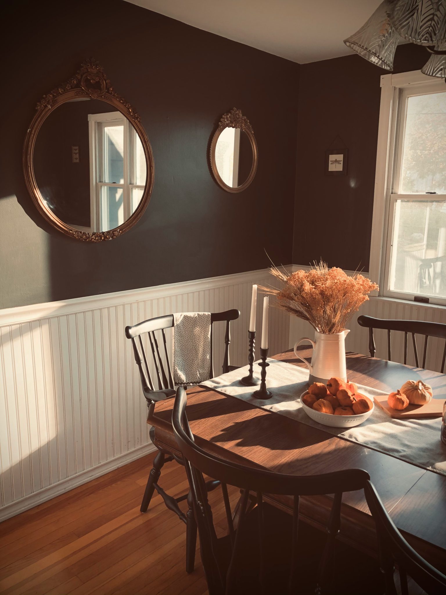 Antique dark brown wooden table surrounded by vintage black chairs in a dark green walled dining room. Ornate vintage gold mirrors hang on the walls. Faux pumpkins and dried foliage decorate the table
