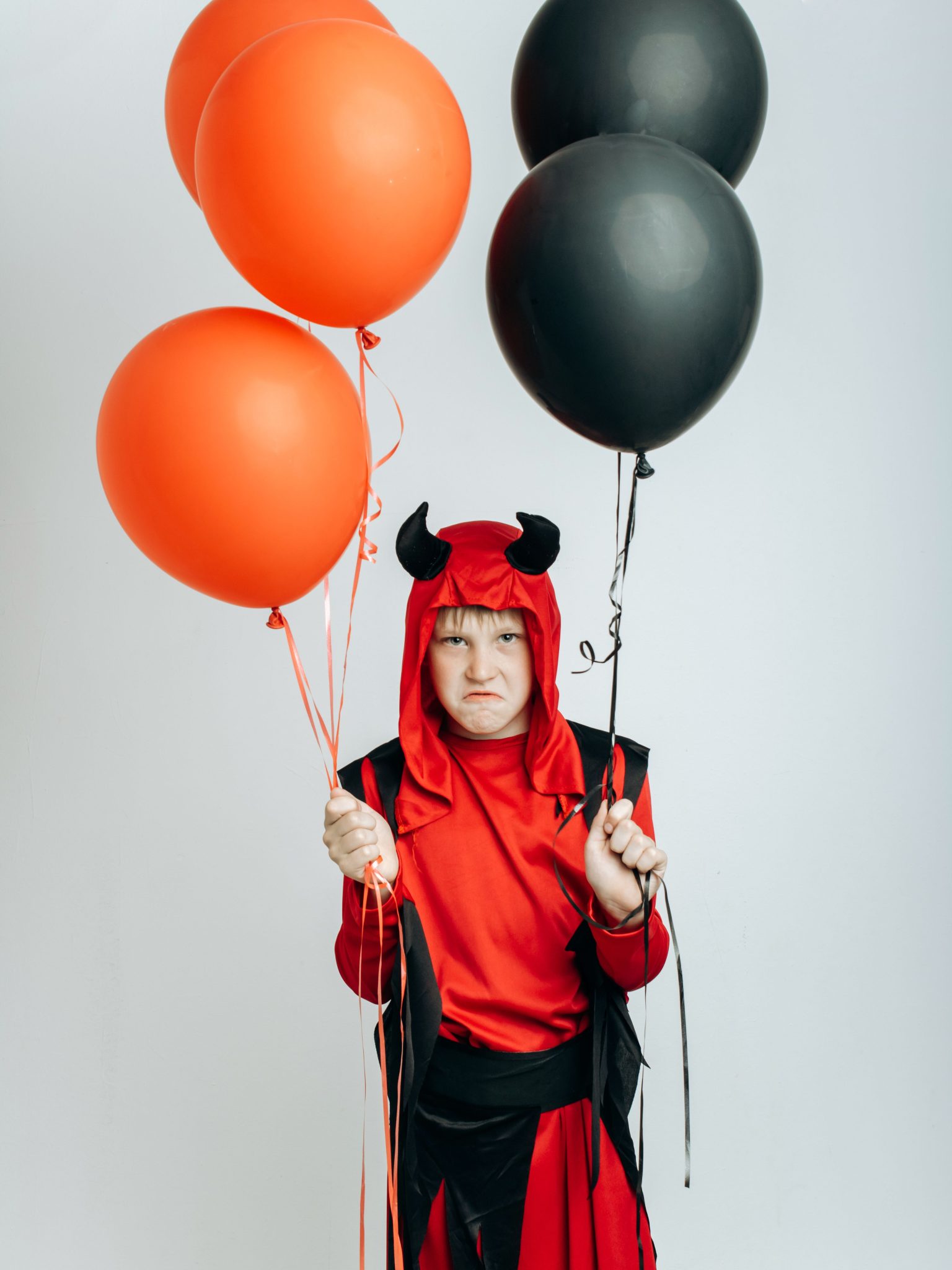 Child in devil costume holding red and black balloons against a plain background.