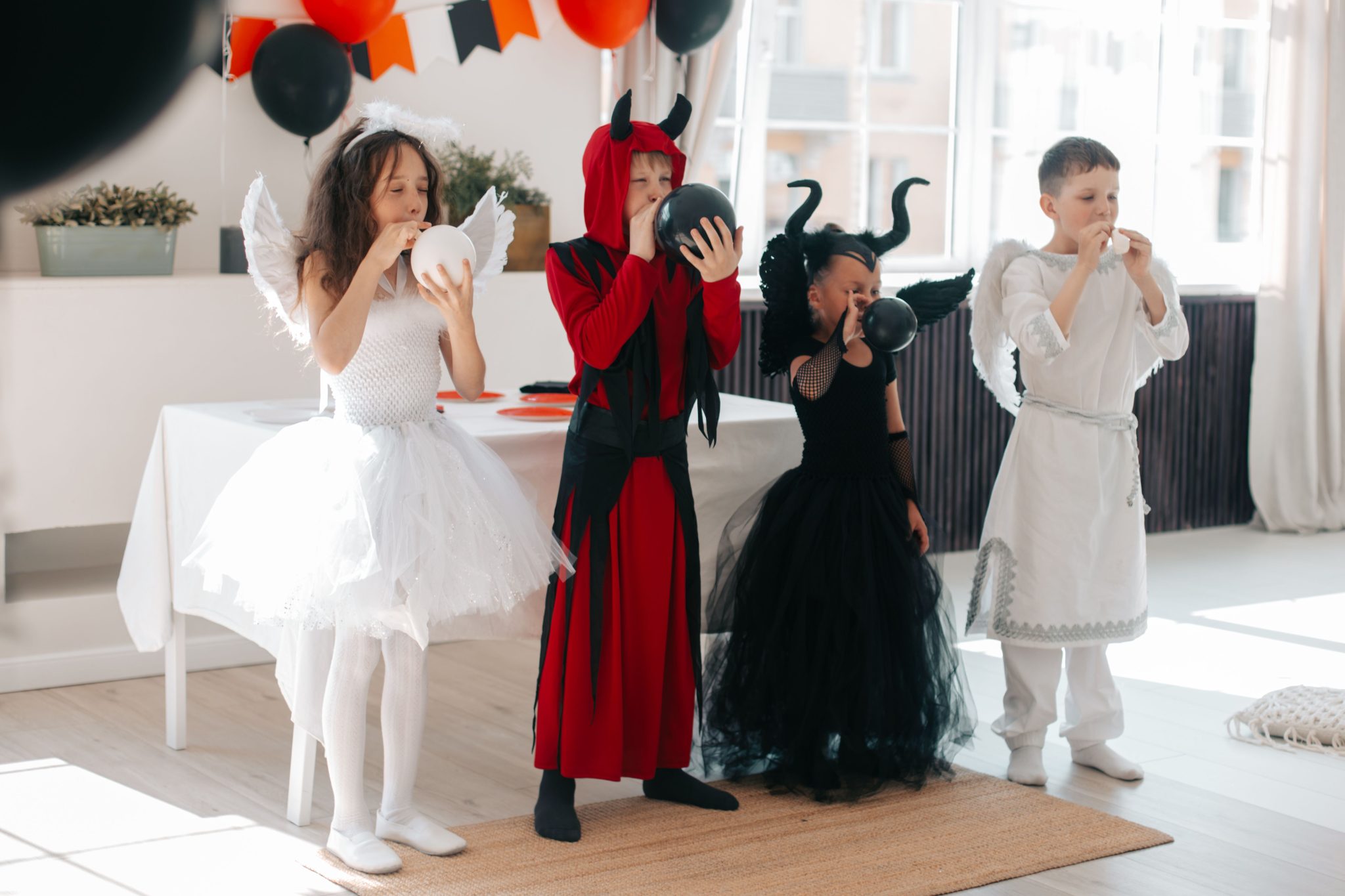 Children in festive Halloween costumes blowing up balloons at a party indoors.