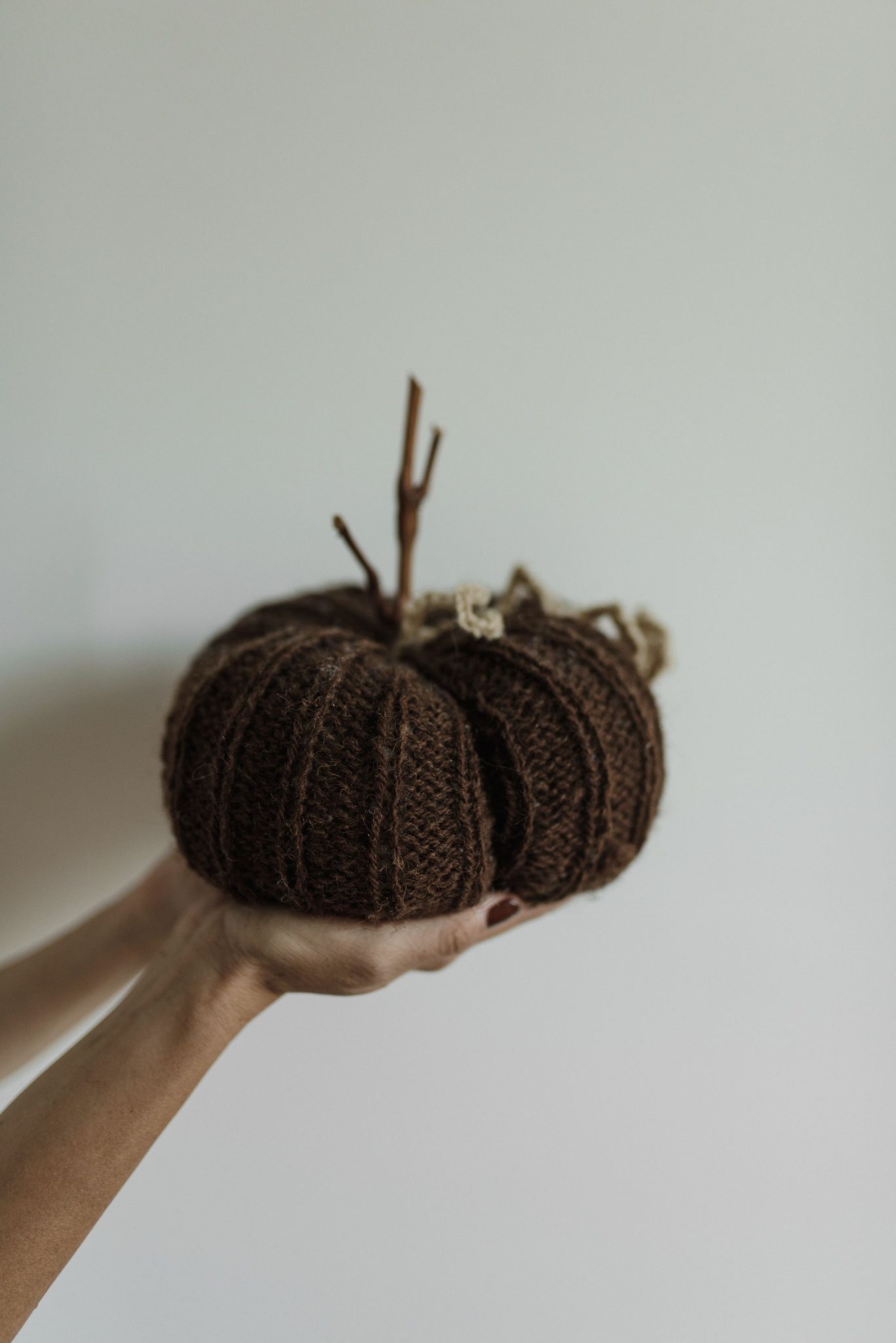 Close-up of a hand holding a knitted brown pumpkin with a stick stem, against a neutral background.