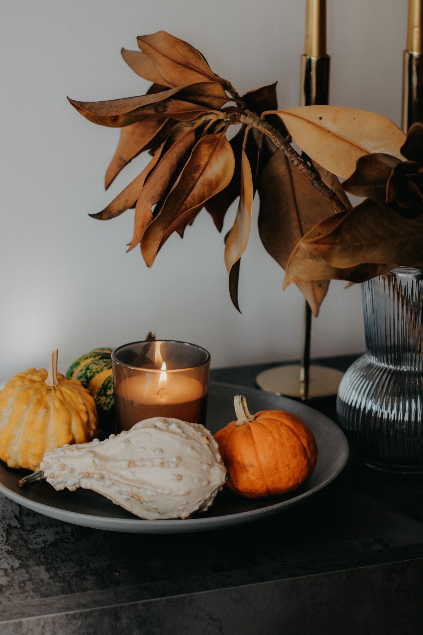 Cozy autumn still life featuring pumpkins and a candle on a decorative plate with dried leaves.