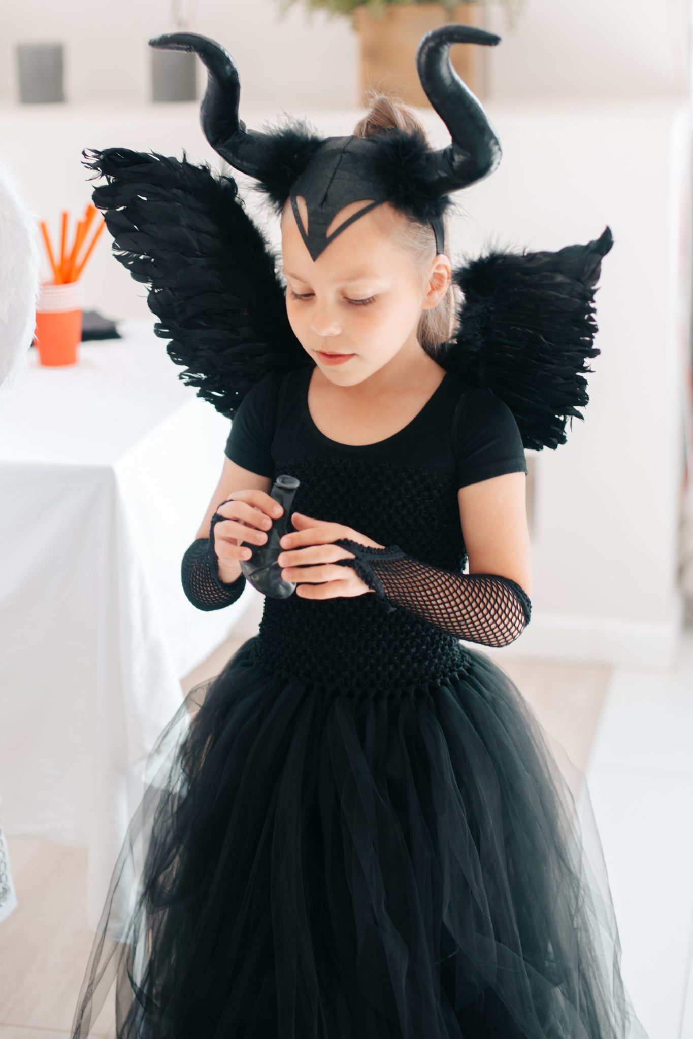 Cute girl in a black devil costume with horns and wings celebrating Halloween indoors.