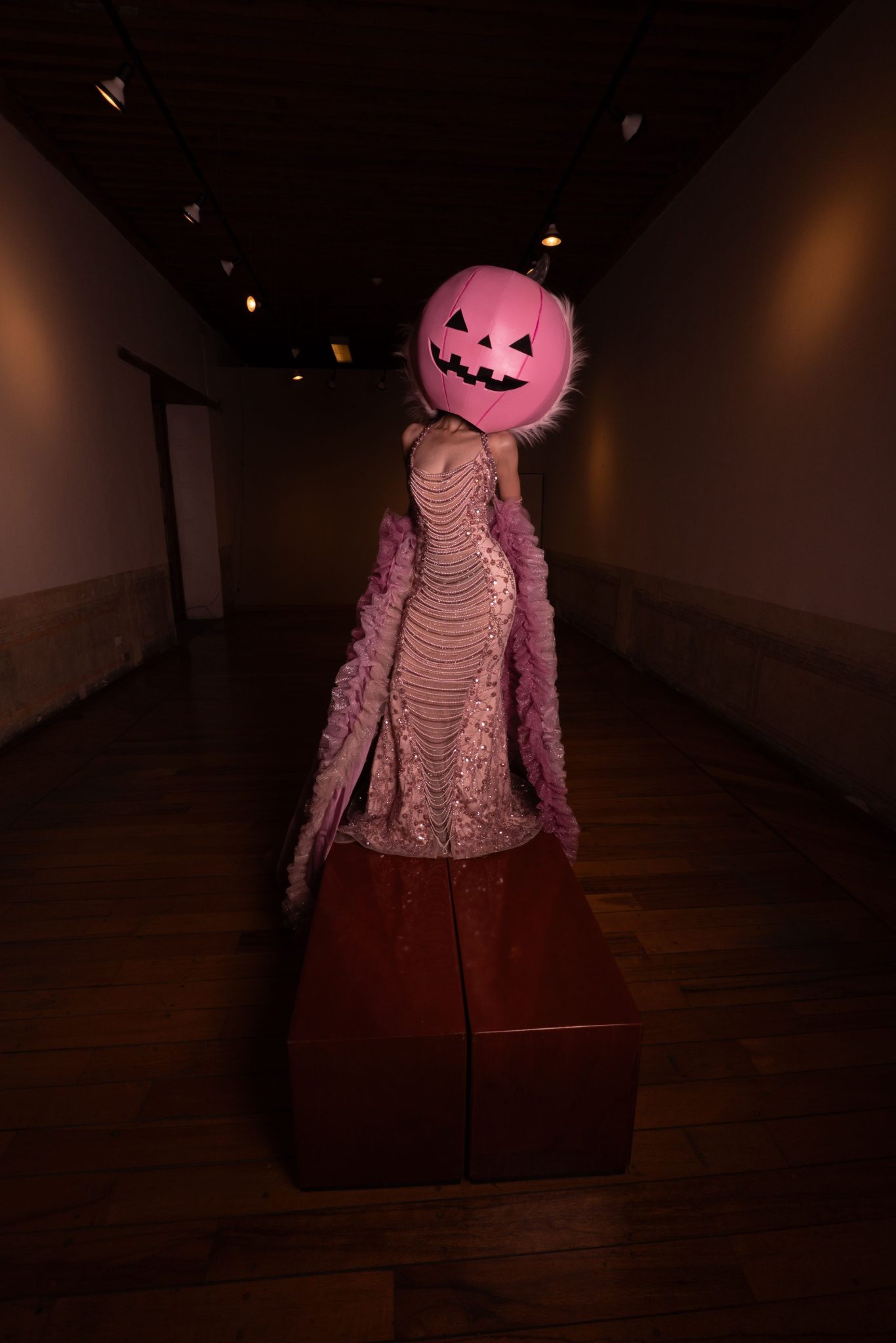 Elegant figure with pink pumpkin head in dimly lit hallway, blending fashion and mystery.