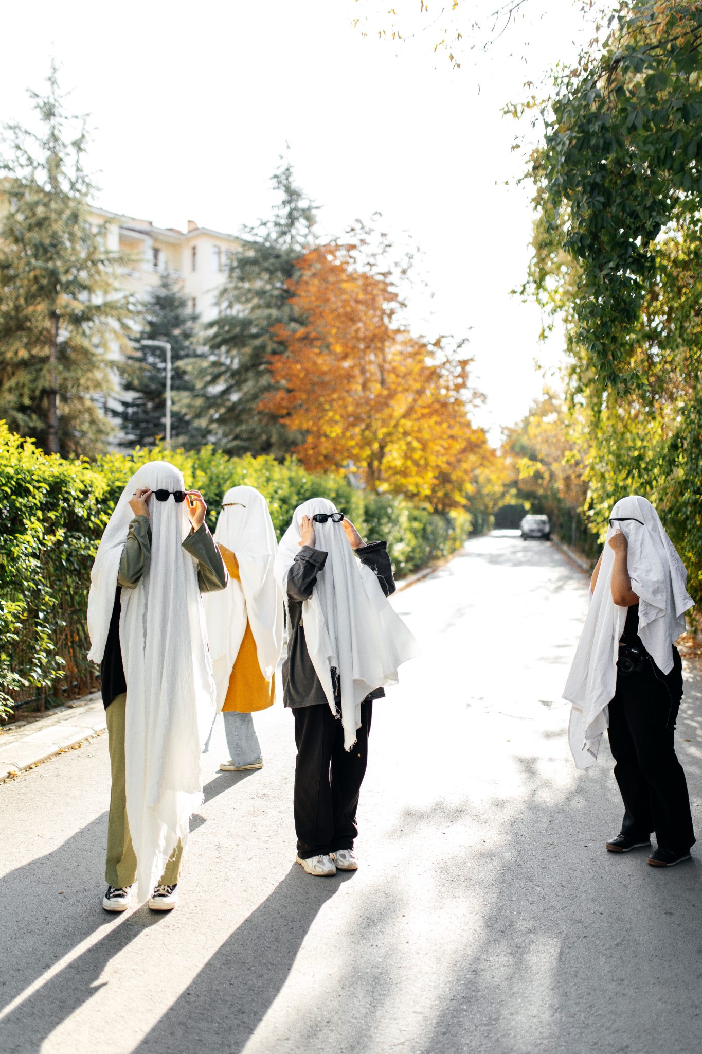 Five people dressed as ghosts standing on a sunlit city street surrounded by autumn foliage.