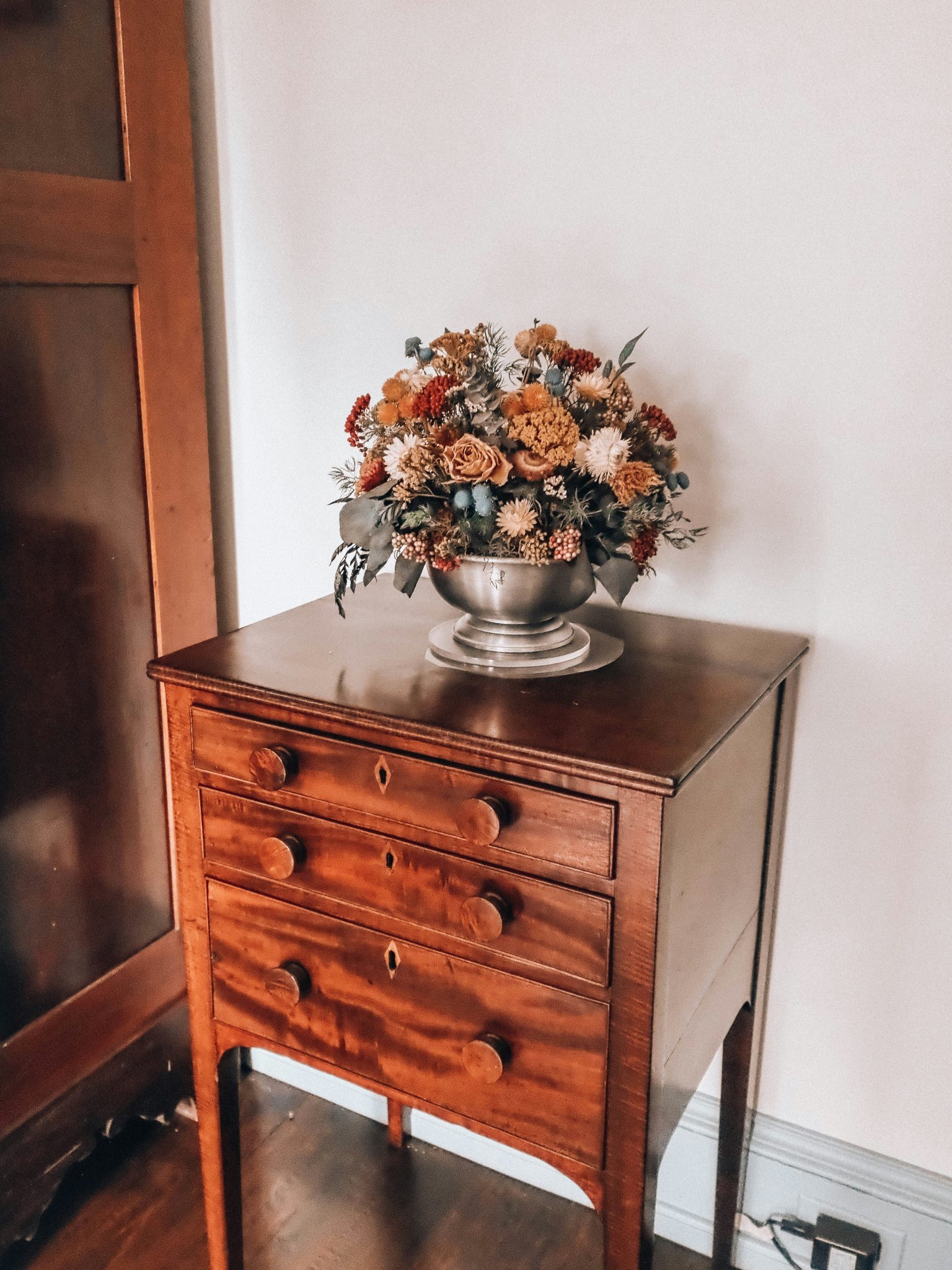 Floral arrangement in silver bowl on antique wooden dresser.