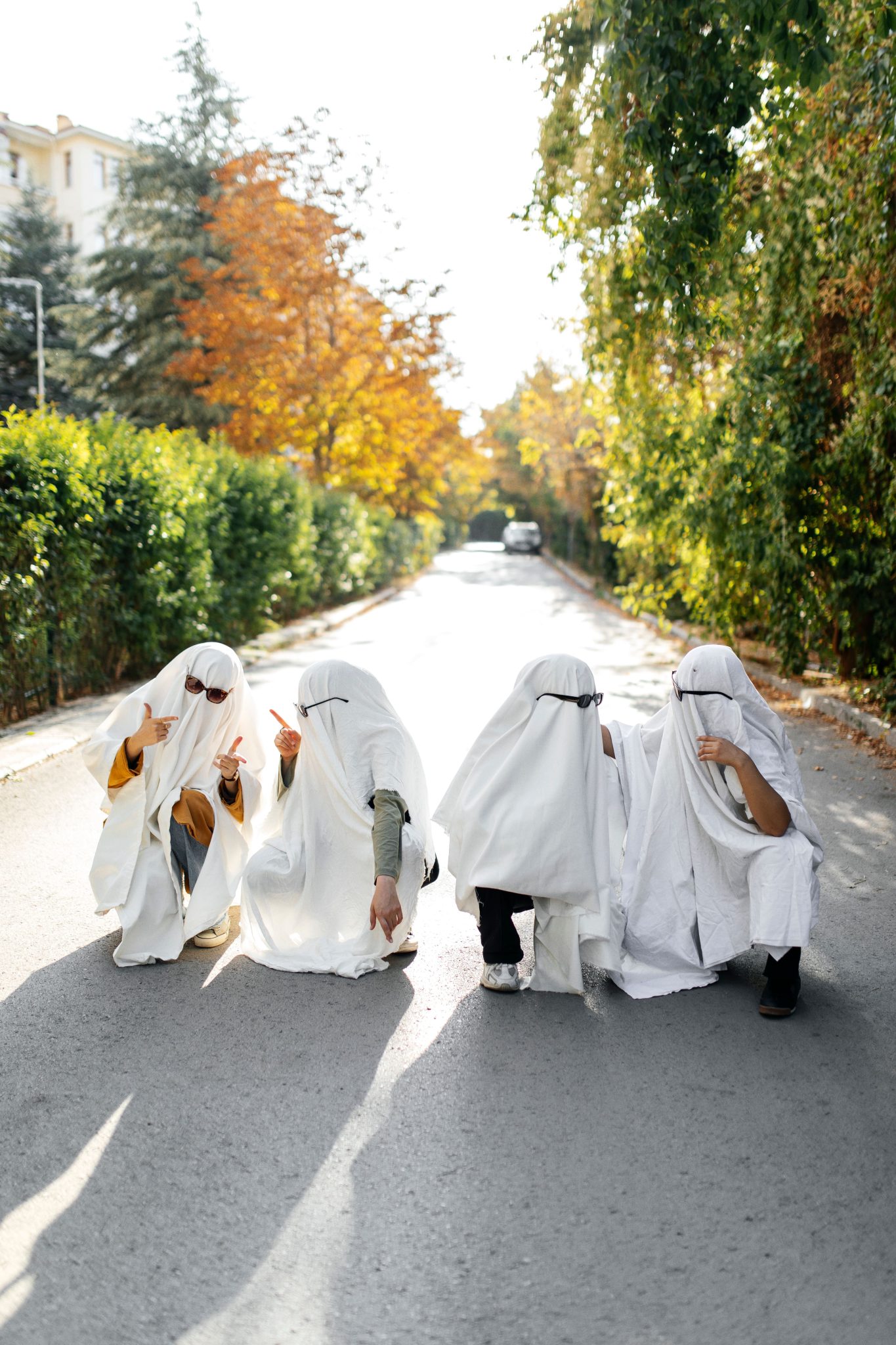 Four individuals in ghost costumes squatting on a sunlit street, capturing a playful Halloween spirit.