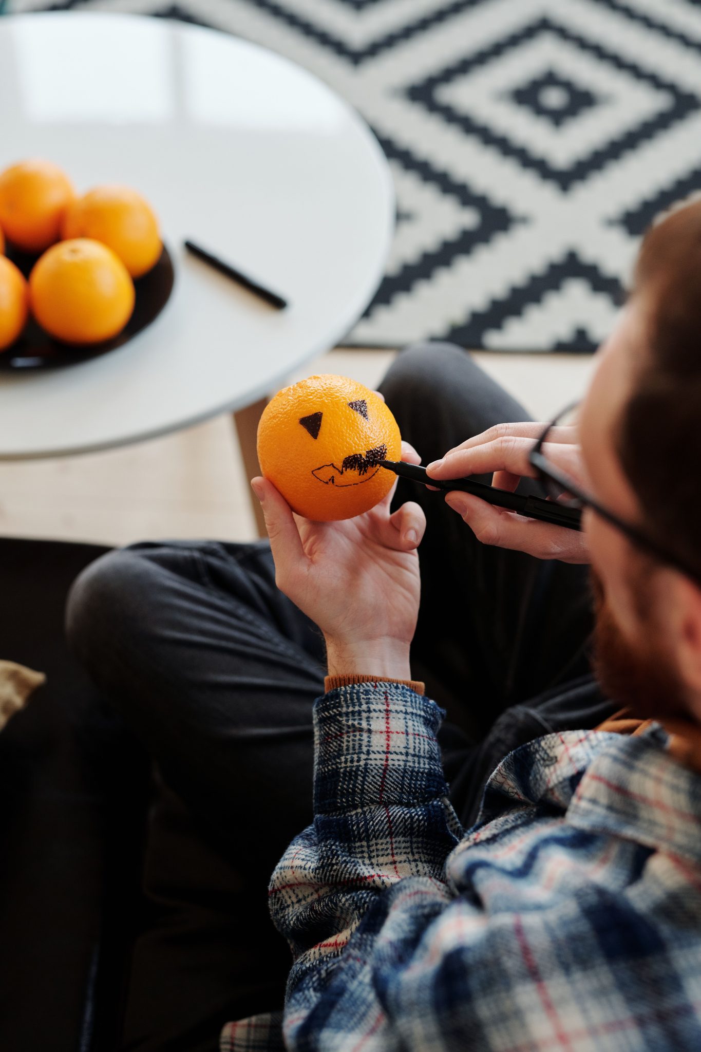Man drawing a jack-o'-lantern face on an orange indoors, enjoying Halloween DIY creativity.