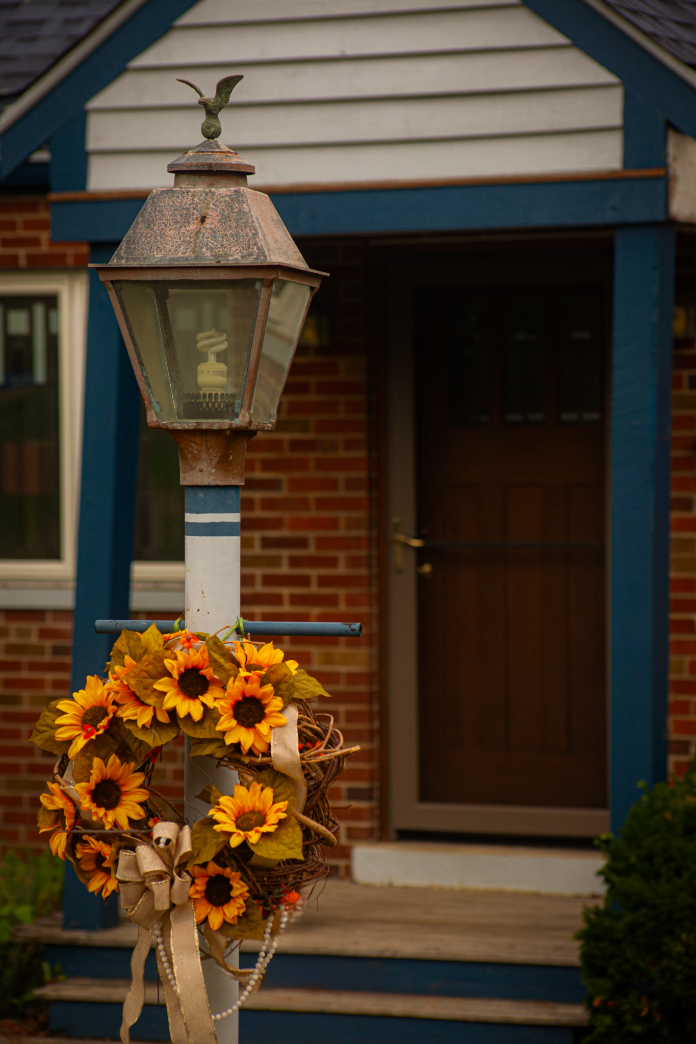 Rustic lamp post adorned with vibrant sunflower wreath in front of a brick house entrance.