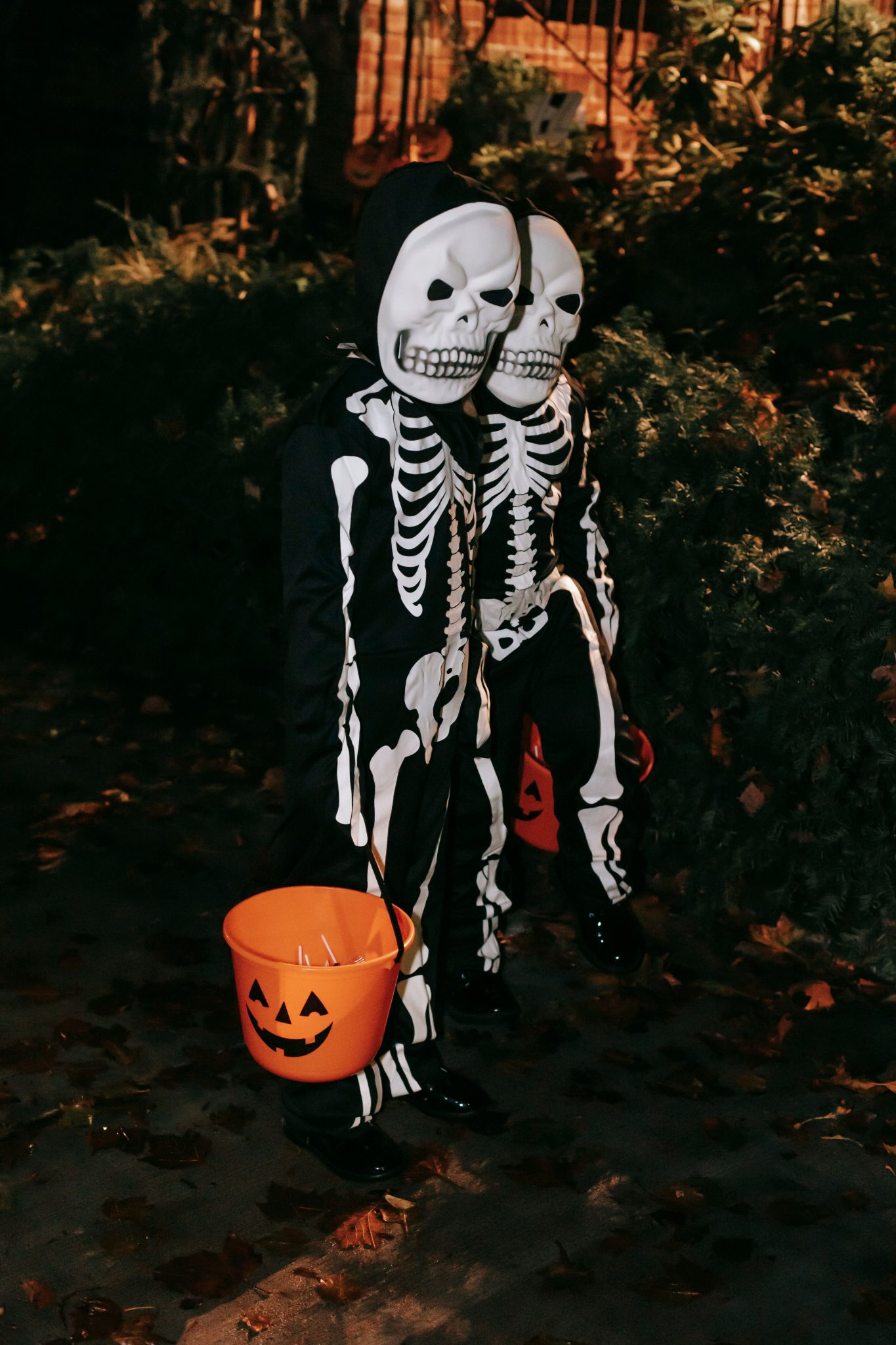 Side view of full length unrecognizable persons in skeleton costumes standing with orange pumpkin buckets with sweets in hands in park at Halloween night