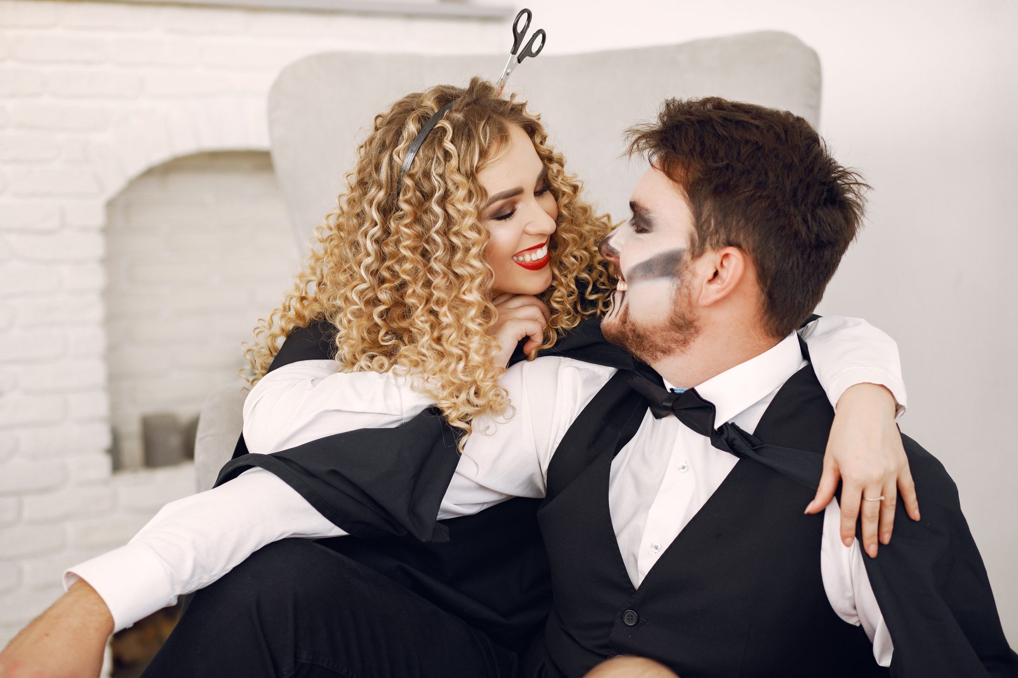 Smiling couple in Halloween costumes with curly hair and face paint, enjoying holiday indoors.