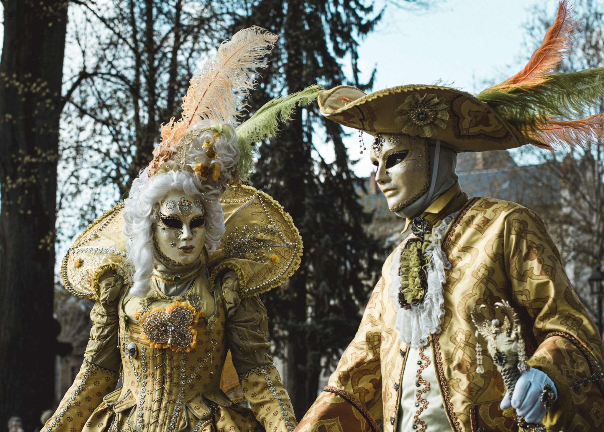 Two adults in ornate Venetian costumes with masks at an outdoor carnival.