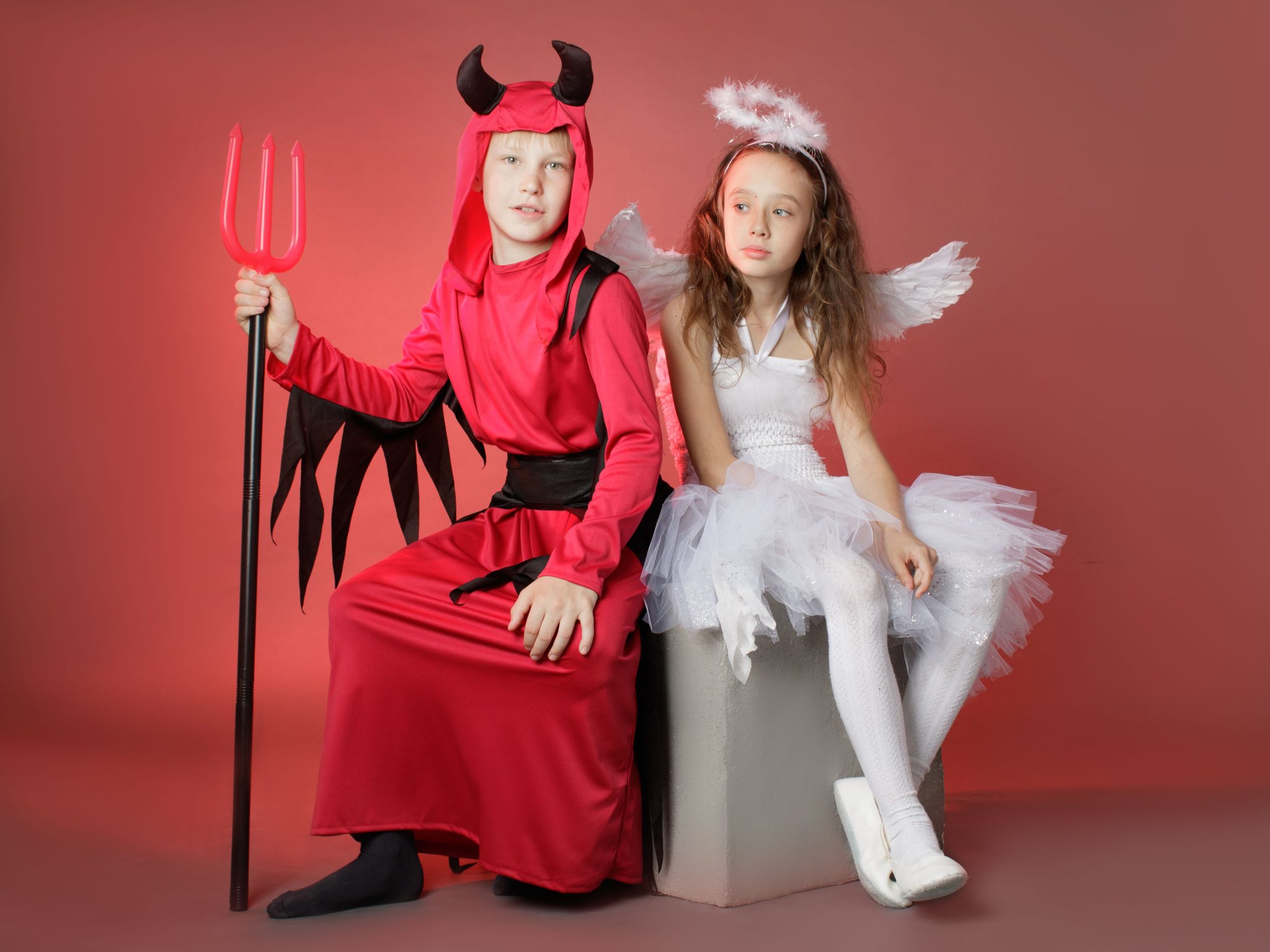 Two children dressed as angel and devil pose in a studio setting for Halloween.