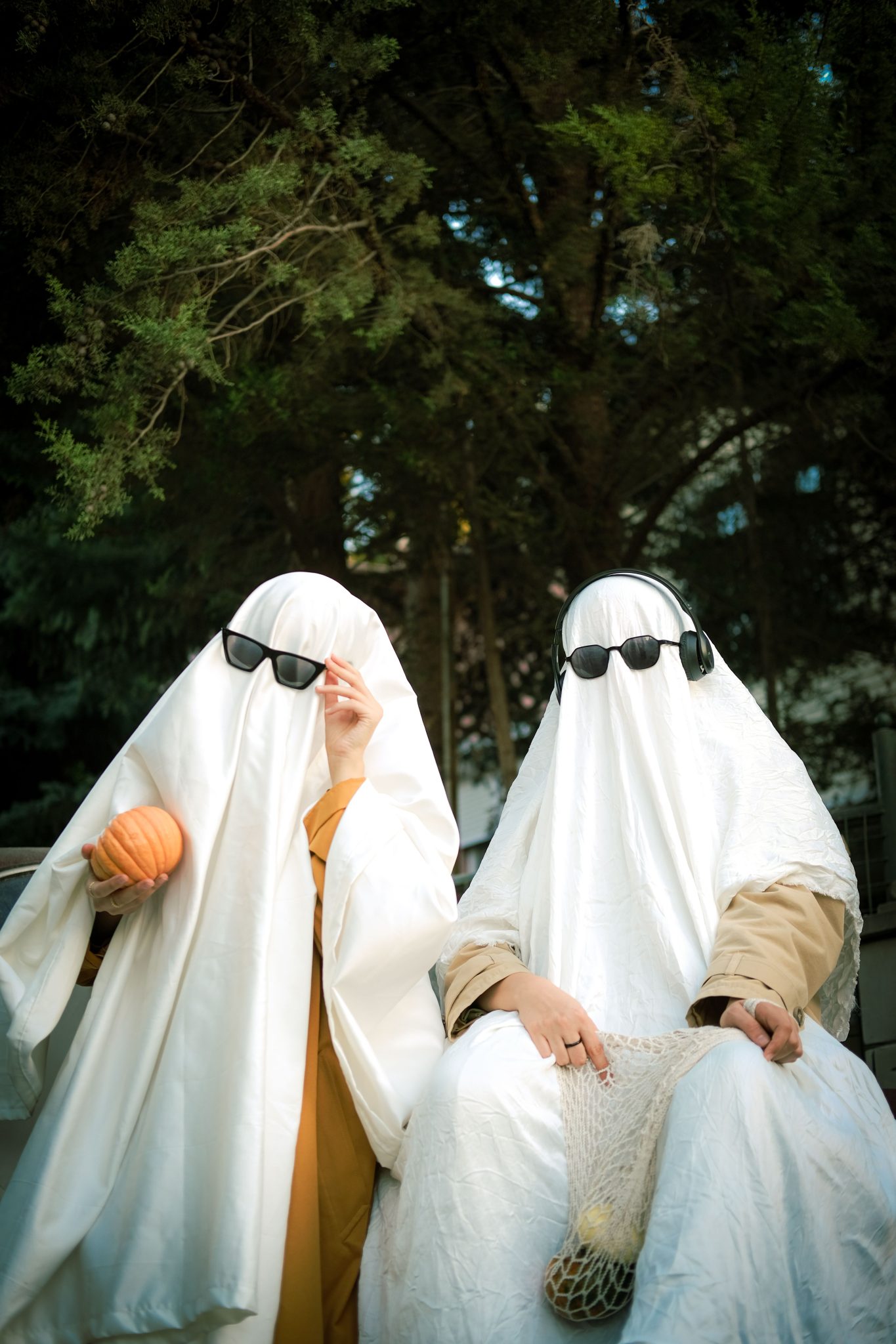 Two people dressed as ghosts with sunglasses sitting outdoors, holding a pumpkin for Halloween fun.