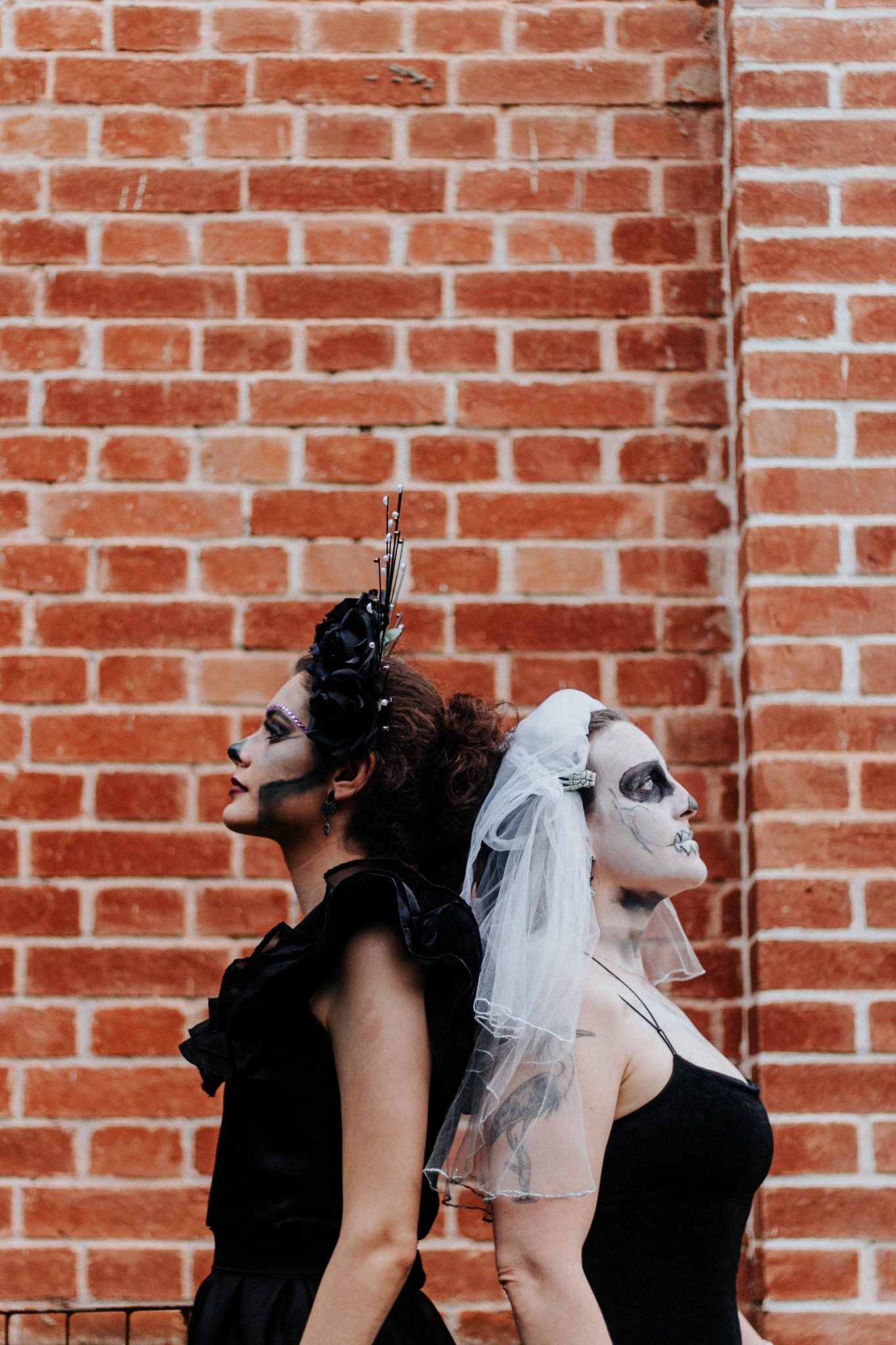 Two women in Day of the Dead costume with intricate makeup against a brick wall.