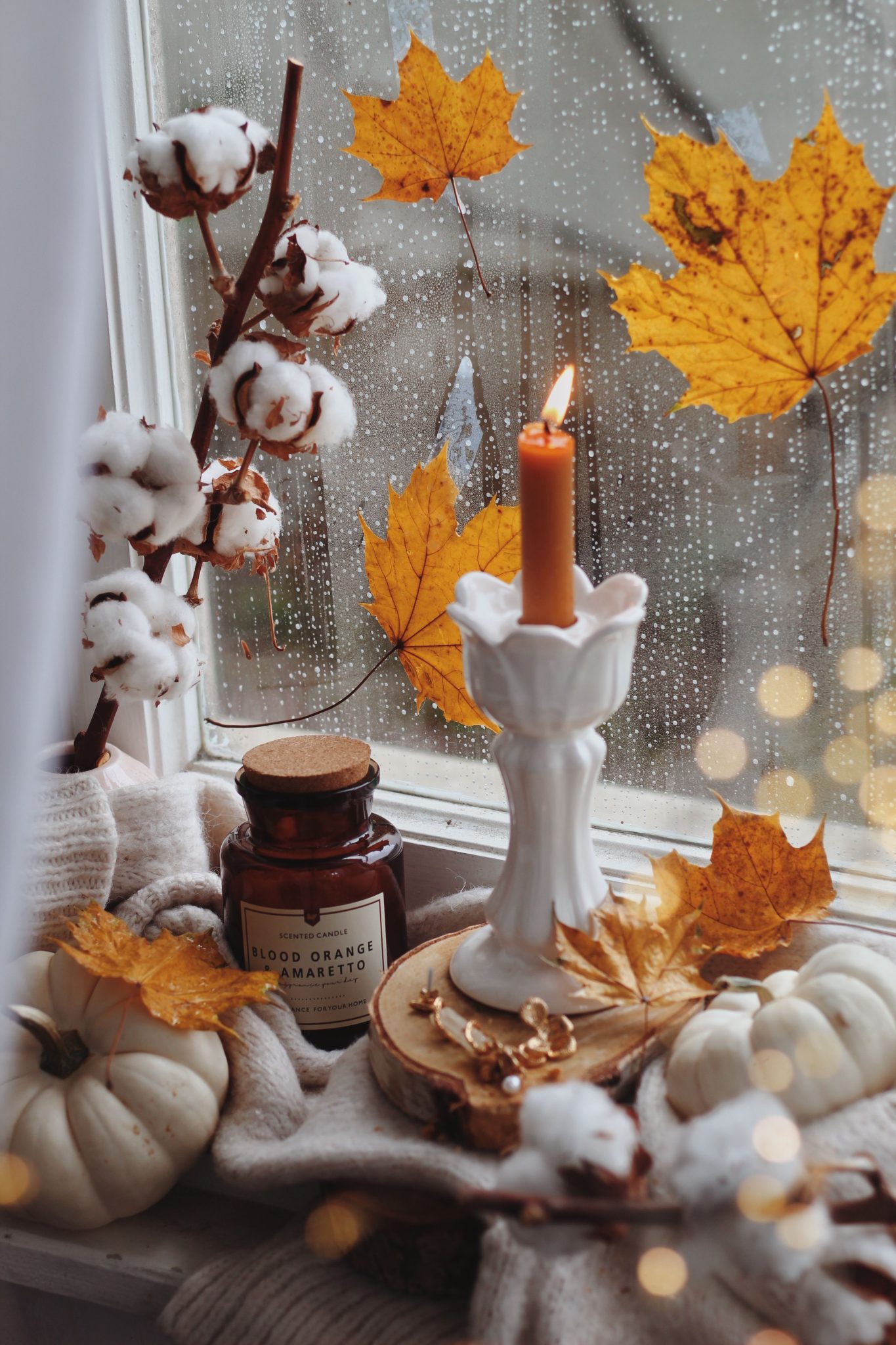 Warm autumn decor with candle, cotton, and maple leaves by a rainy window.