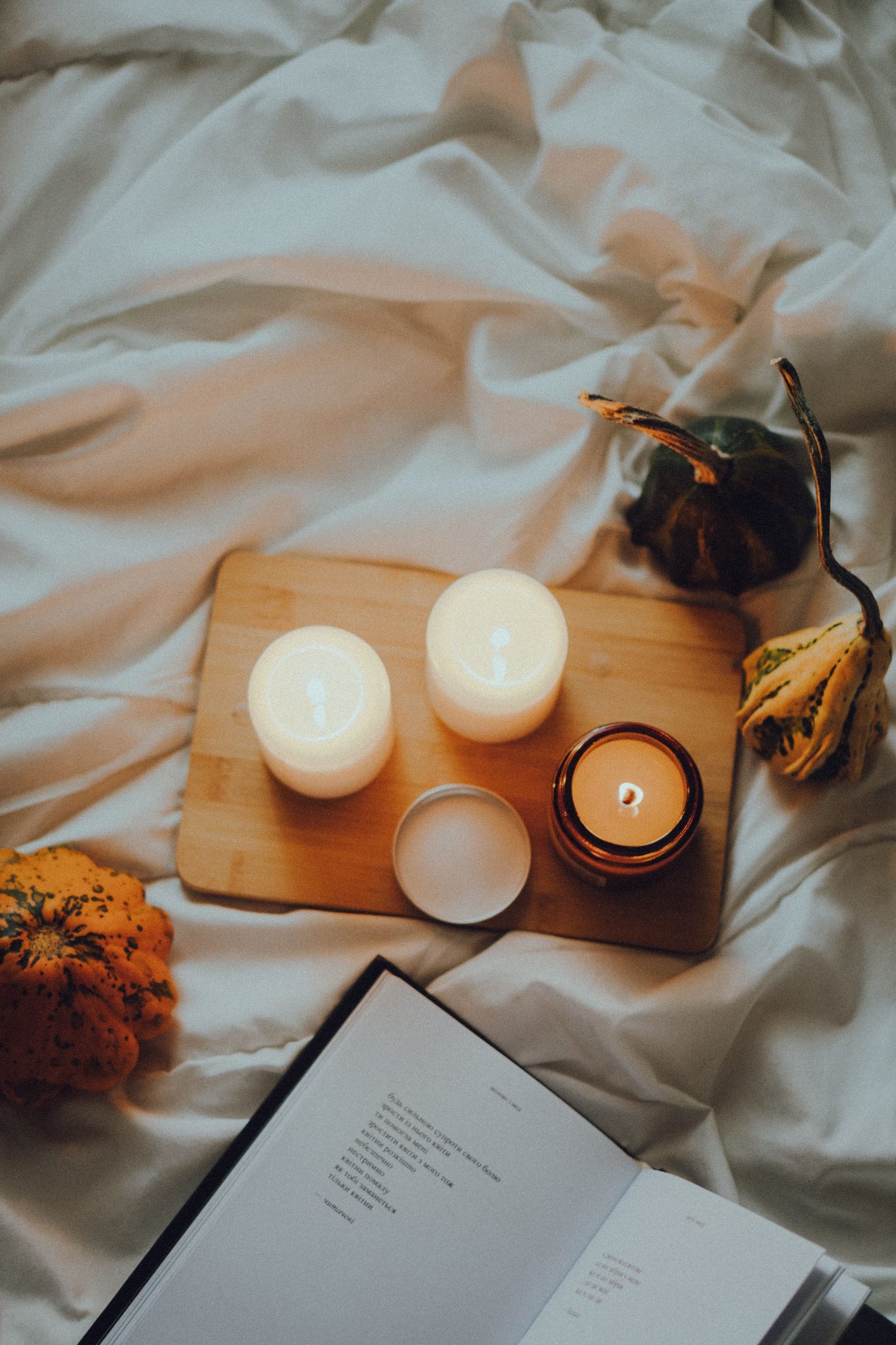 Warm cozy scene with lit candles on a wooden board, surrounded by gourds and an open book on soft fabric.