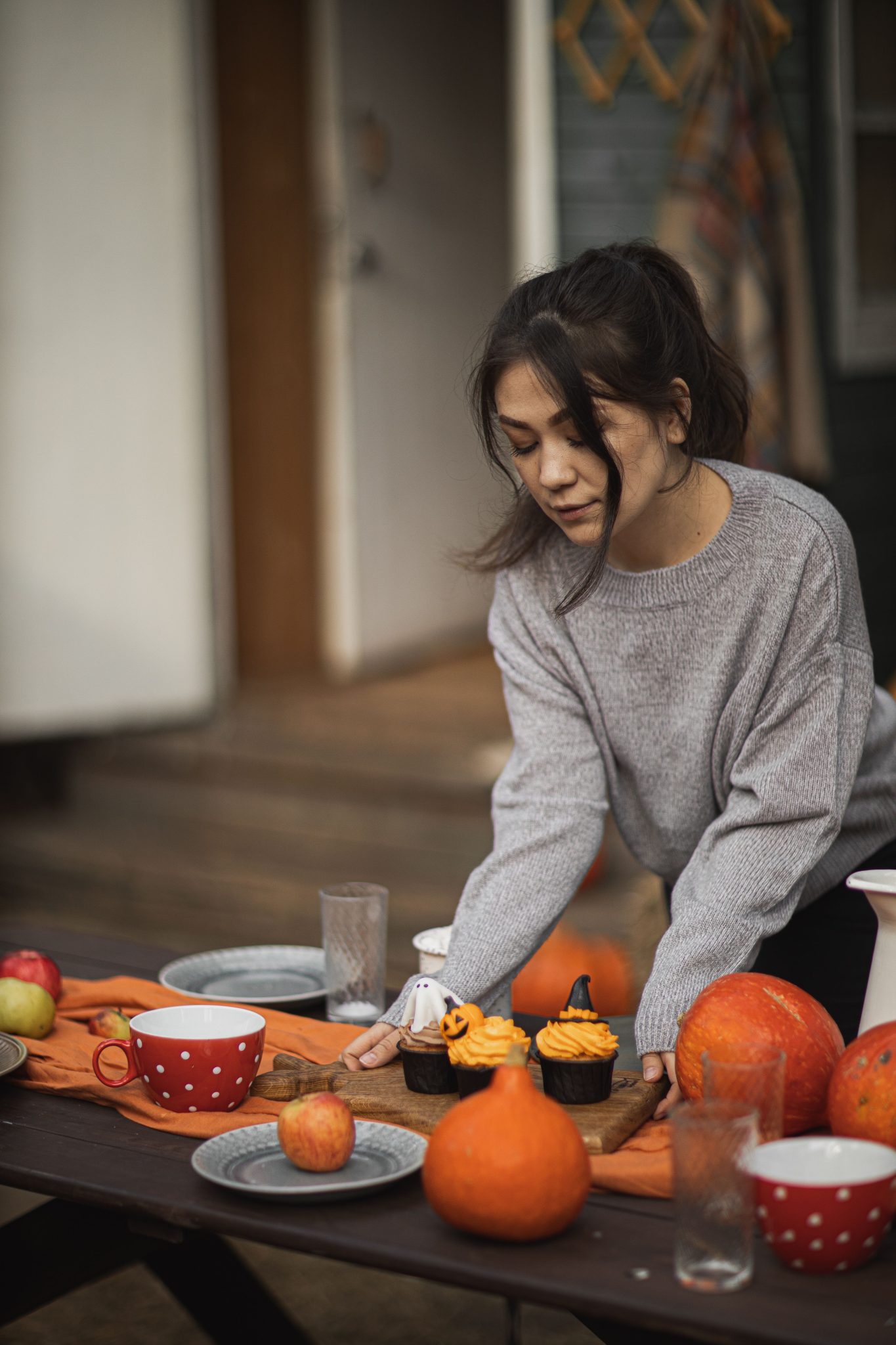 Woman setting a fall-themed table with pumpkins and cupcakes outdoors.