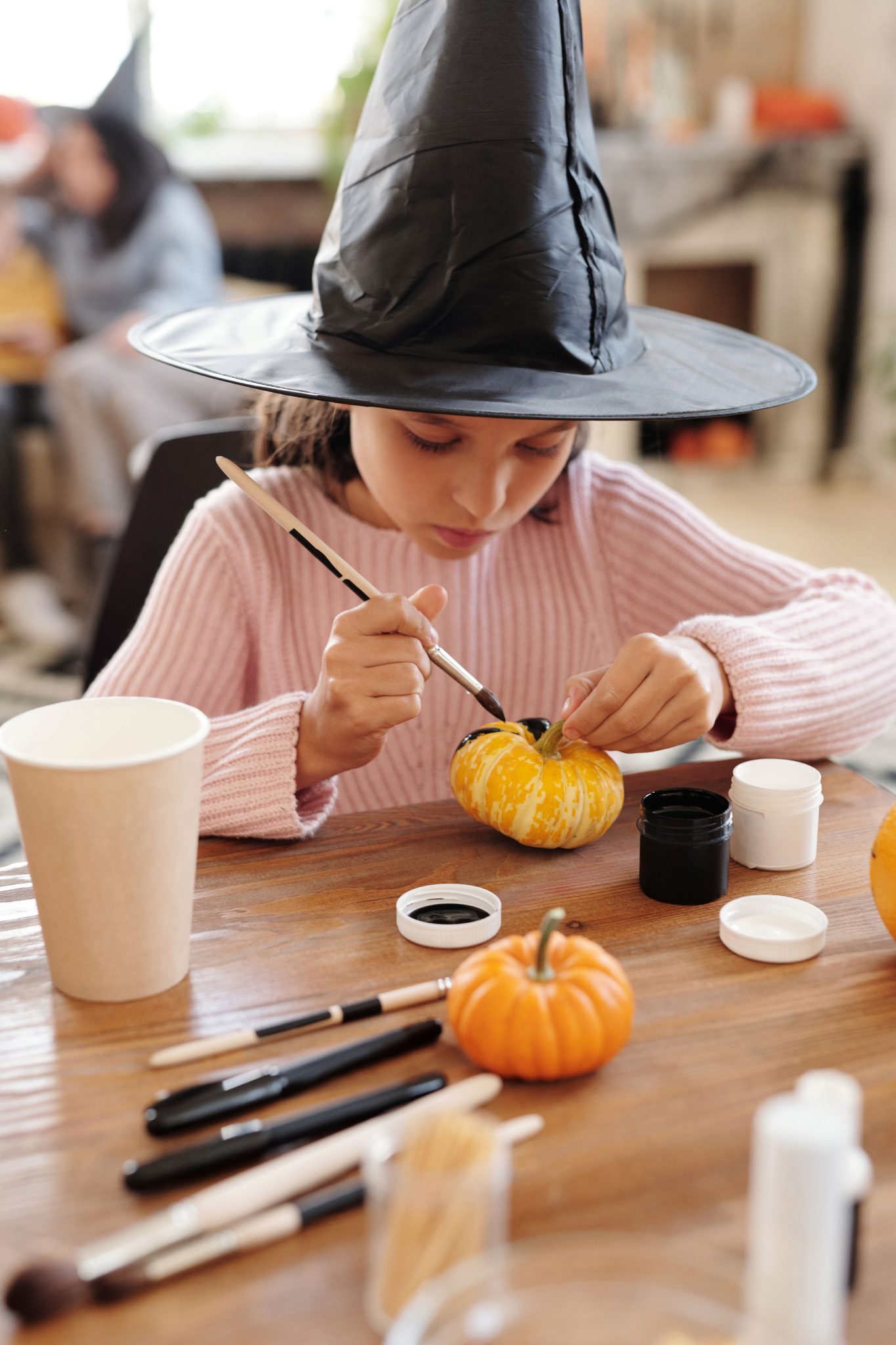 Young girl painting a pumpkin for Halloween at home, wearing a witch hat.