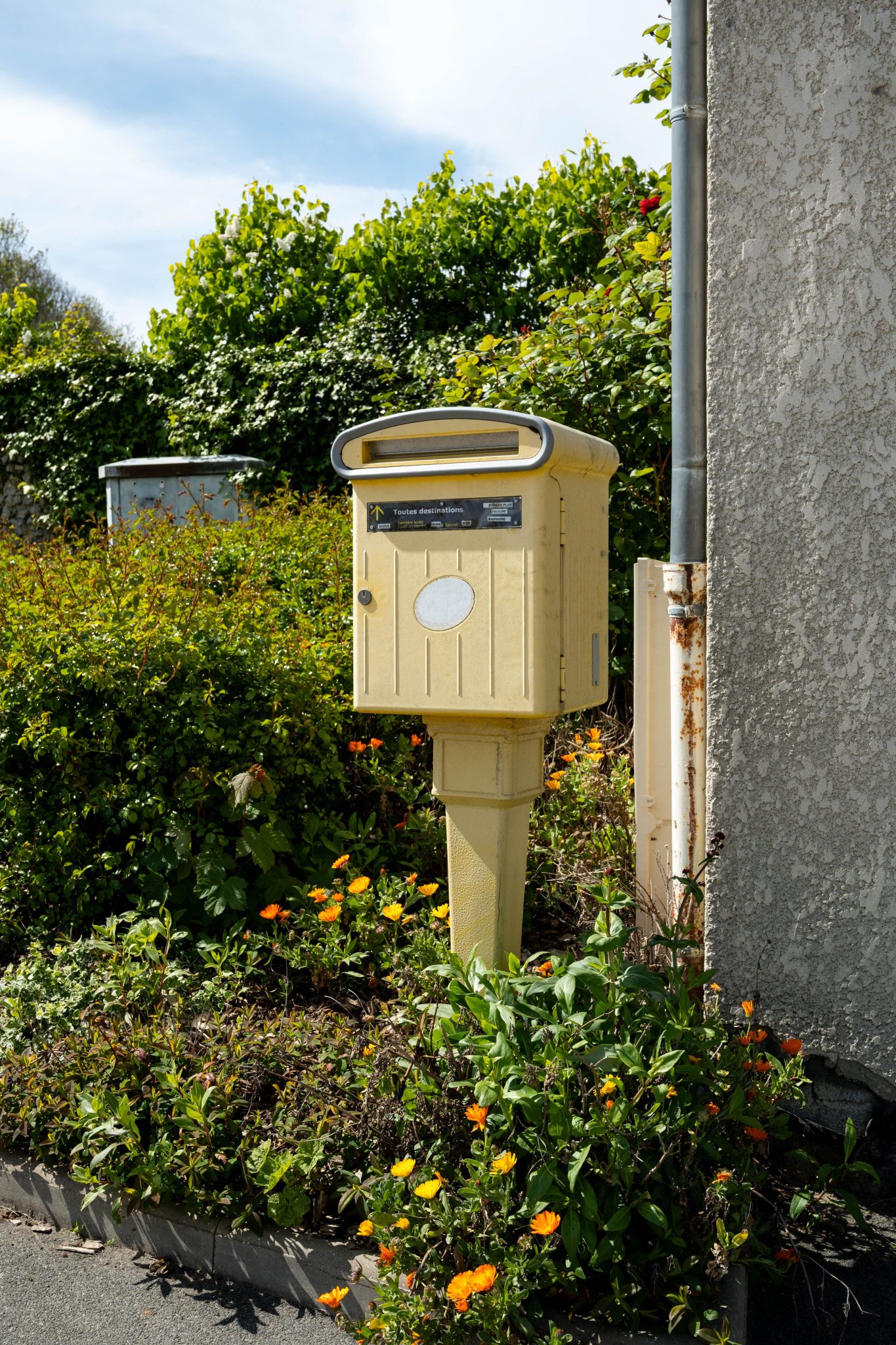 A classic yellow French mailbox set against a vibrant garden backdrop in Escalles, Hauts-de-France.