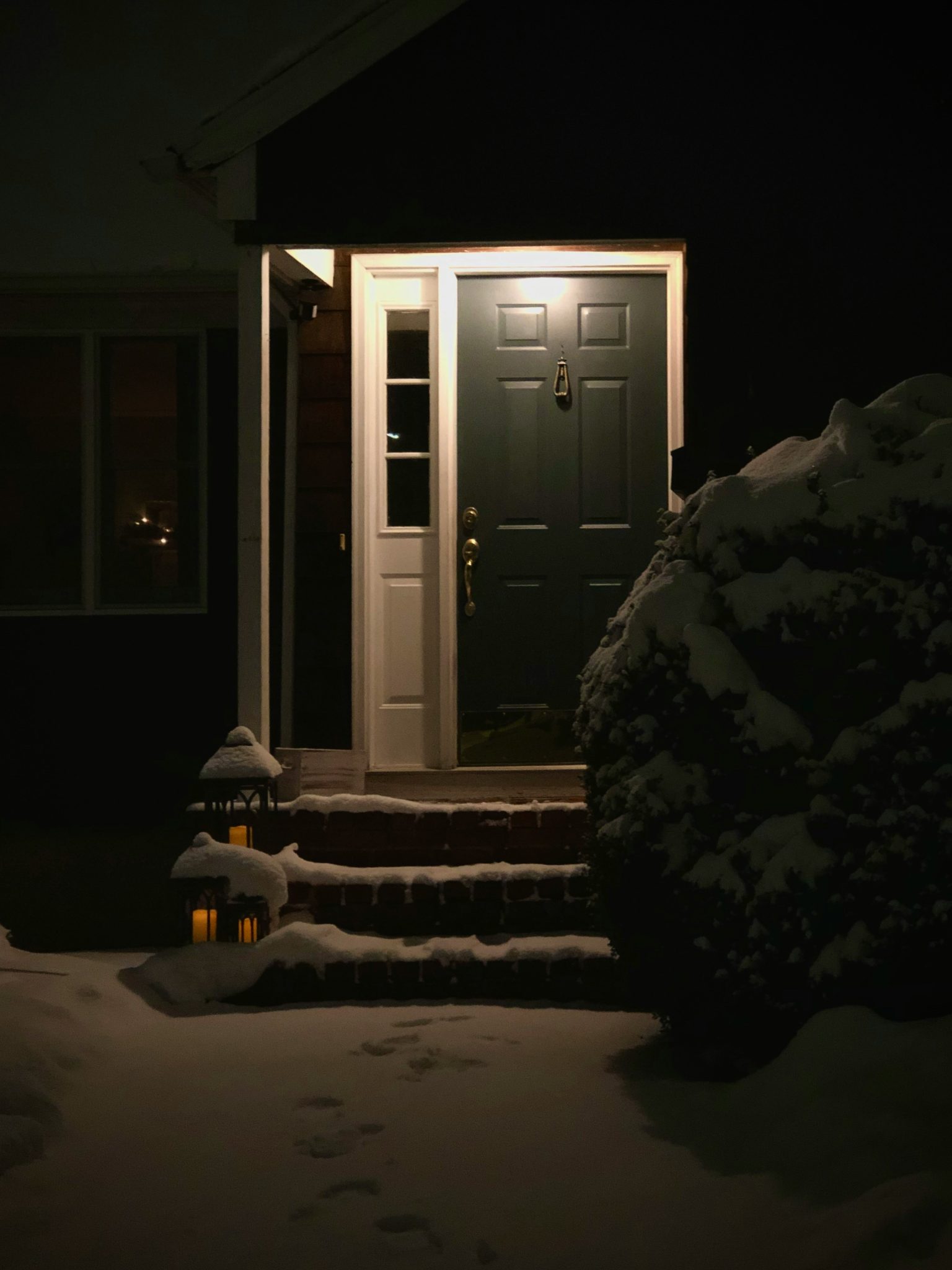 A dark colored front door with an old brass door knocker is illuminated in the darkness of a fresh snowfall.