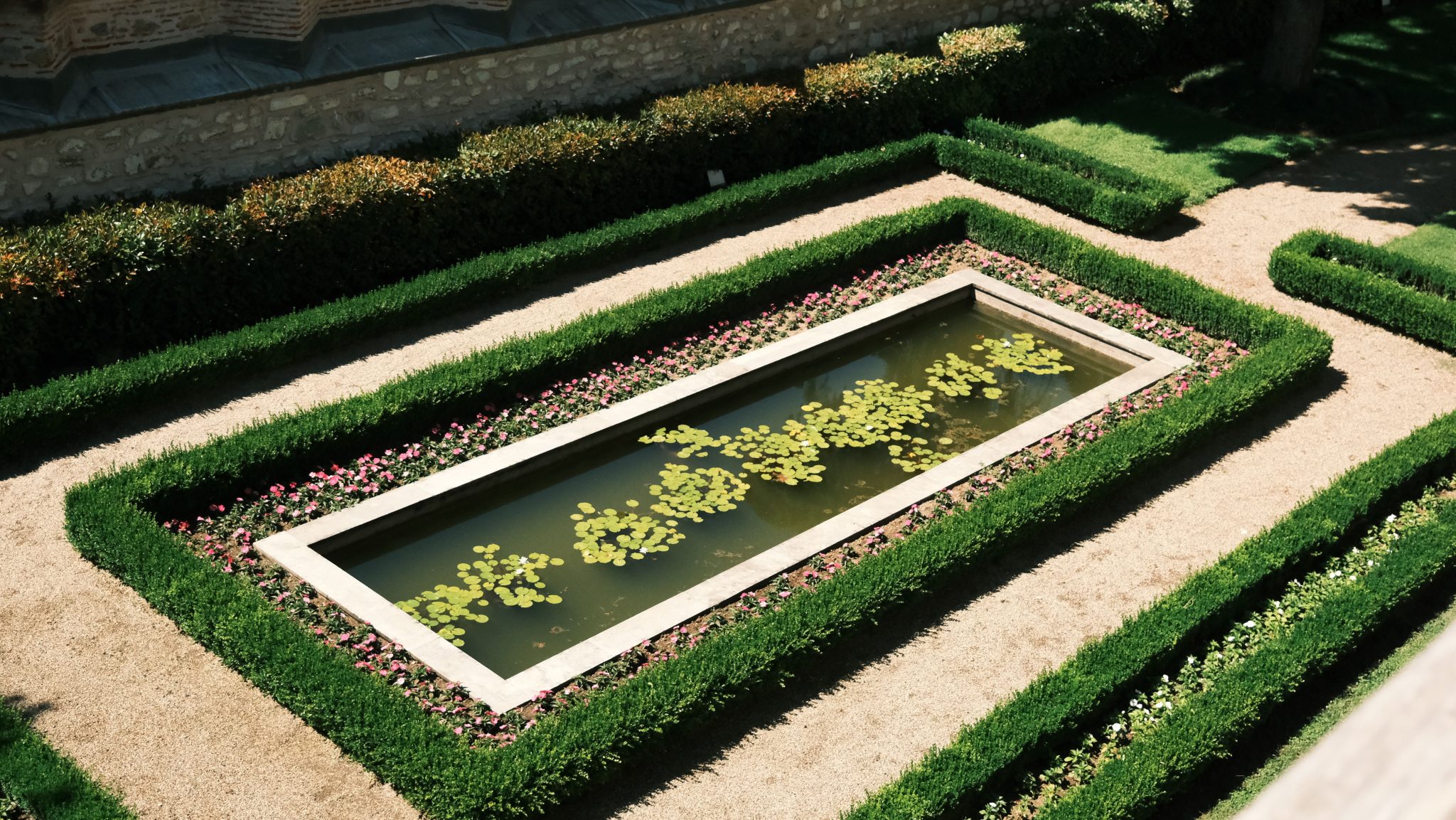 A top view of an elegant rectangular pond surrounded by symmetrical hedges and flowers, in bright daylight.