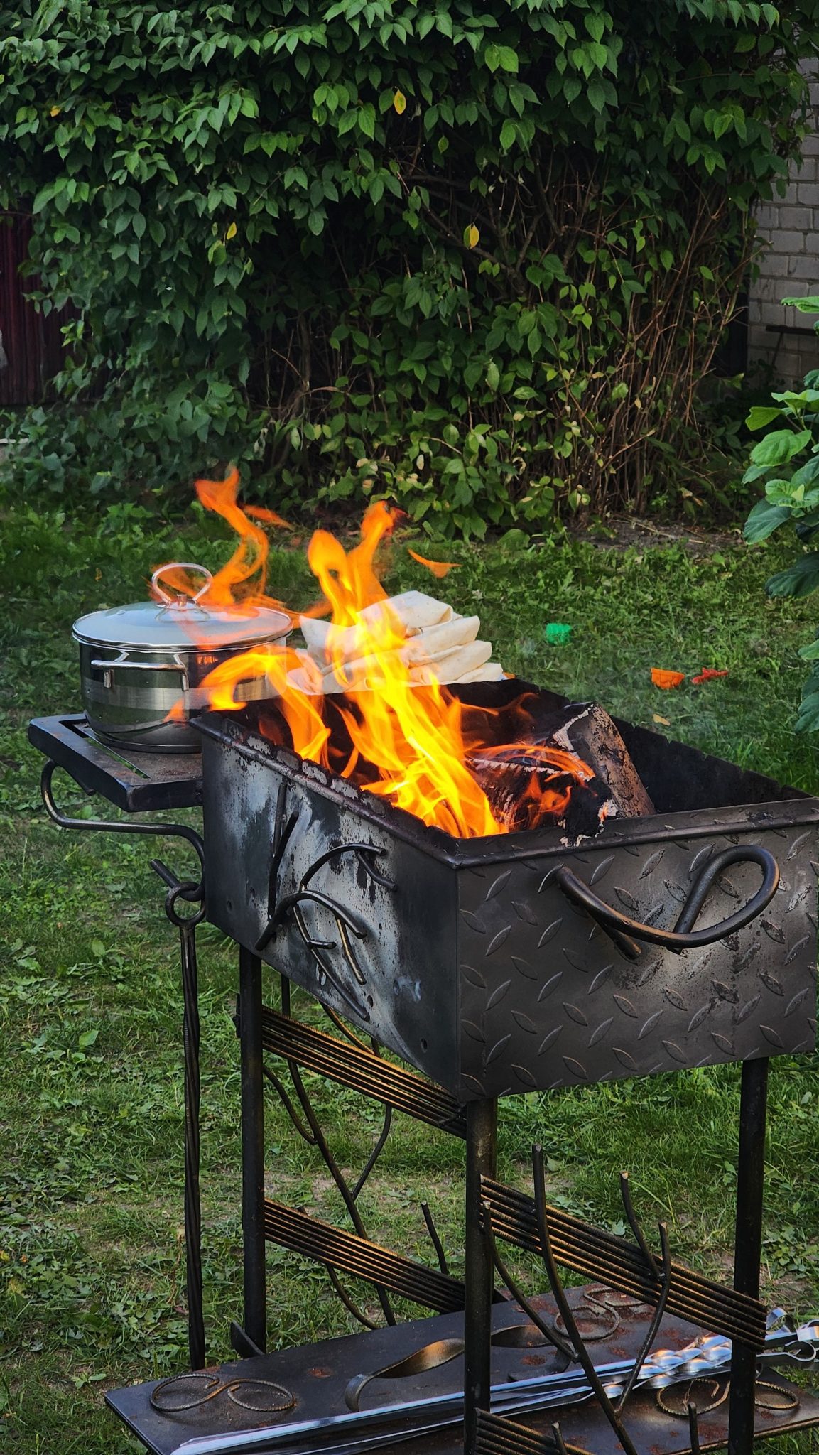 An outdoor barbecue grill with bright flames and a cooking pot in a lush garden setting.