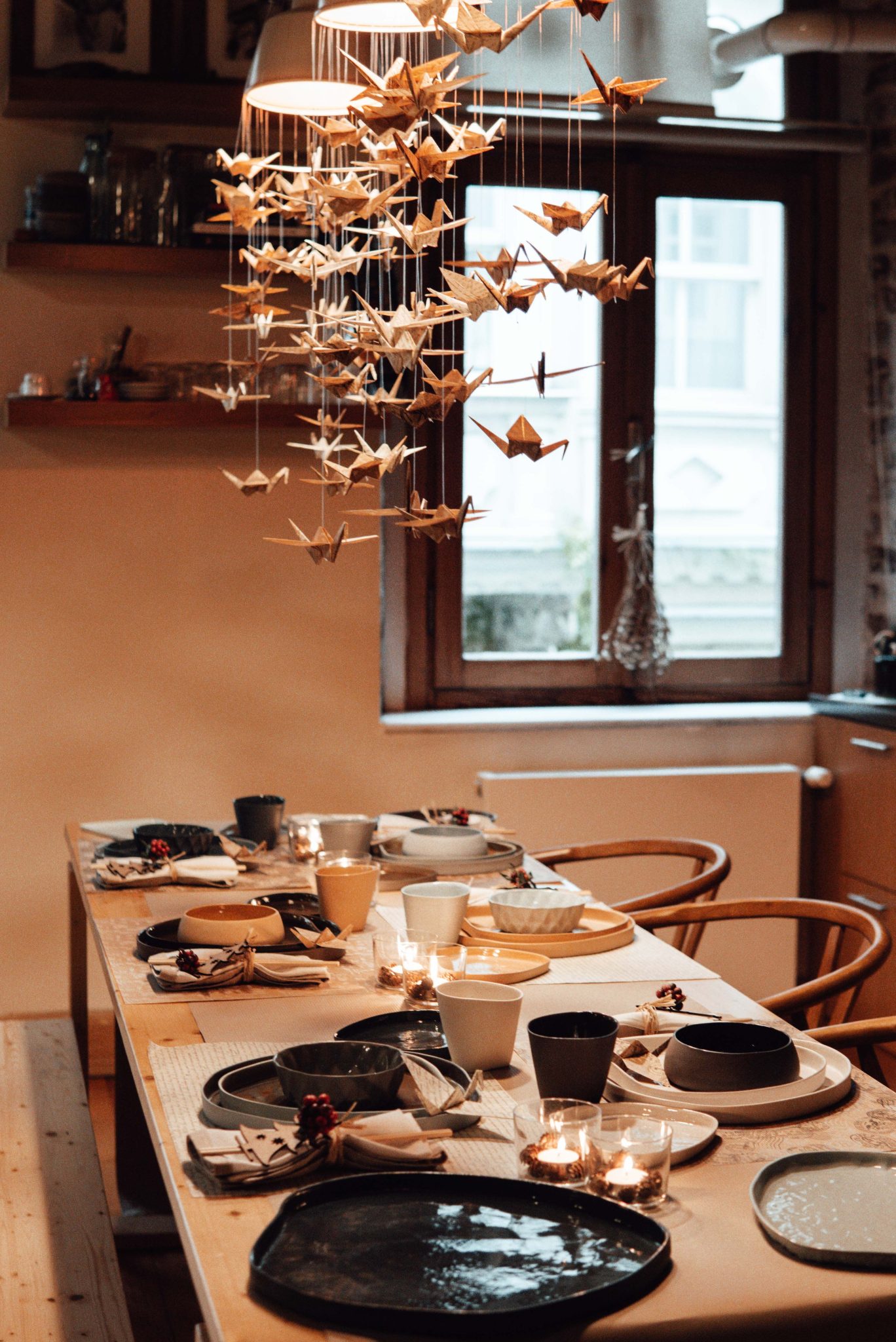 Dining table served with various dishware and burning candles in stylish kitchen with window and hanging paper cranes at home