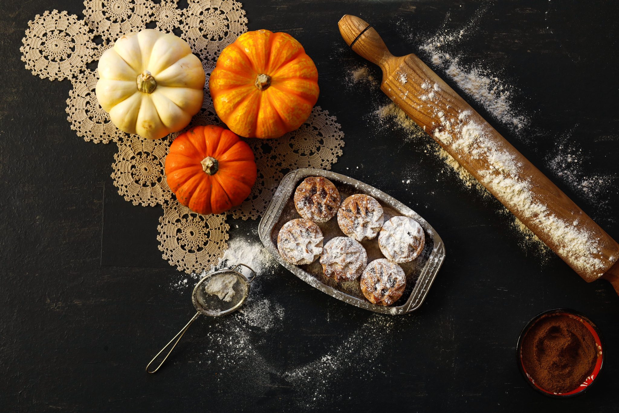 Flat lay of pumpkins, pastries, rolling pin, and sieve on a dark surface.